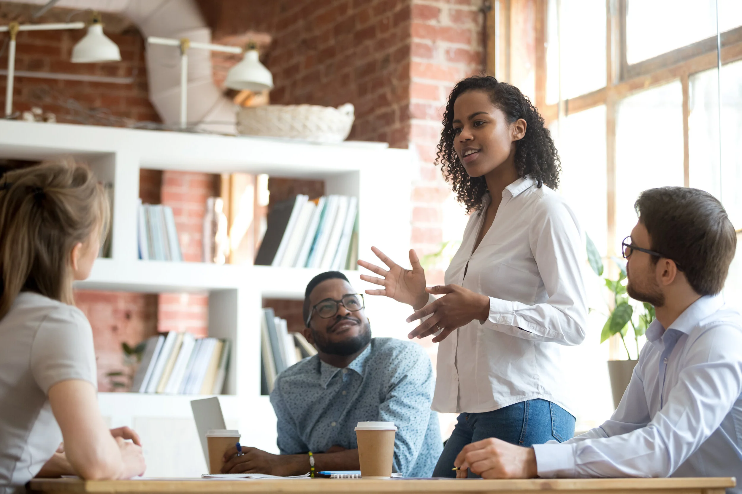 Woman standing at table speaking in front of three other coworkers