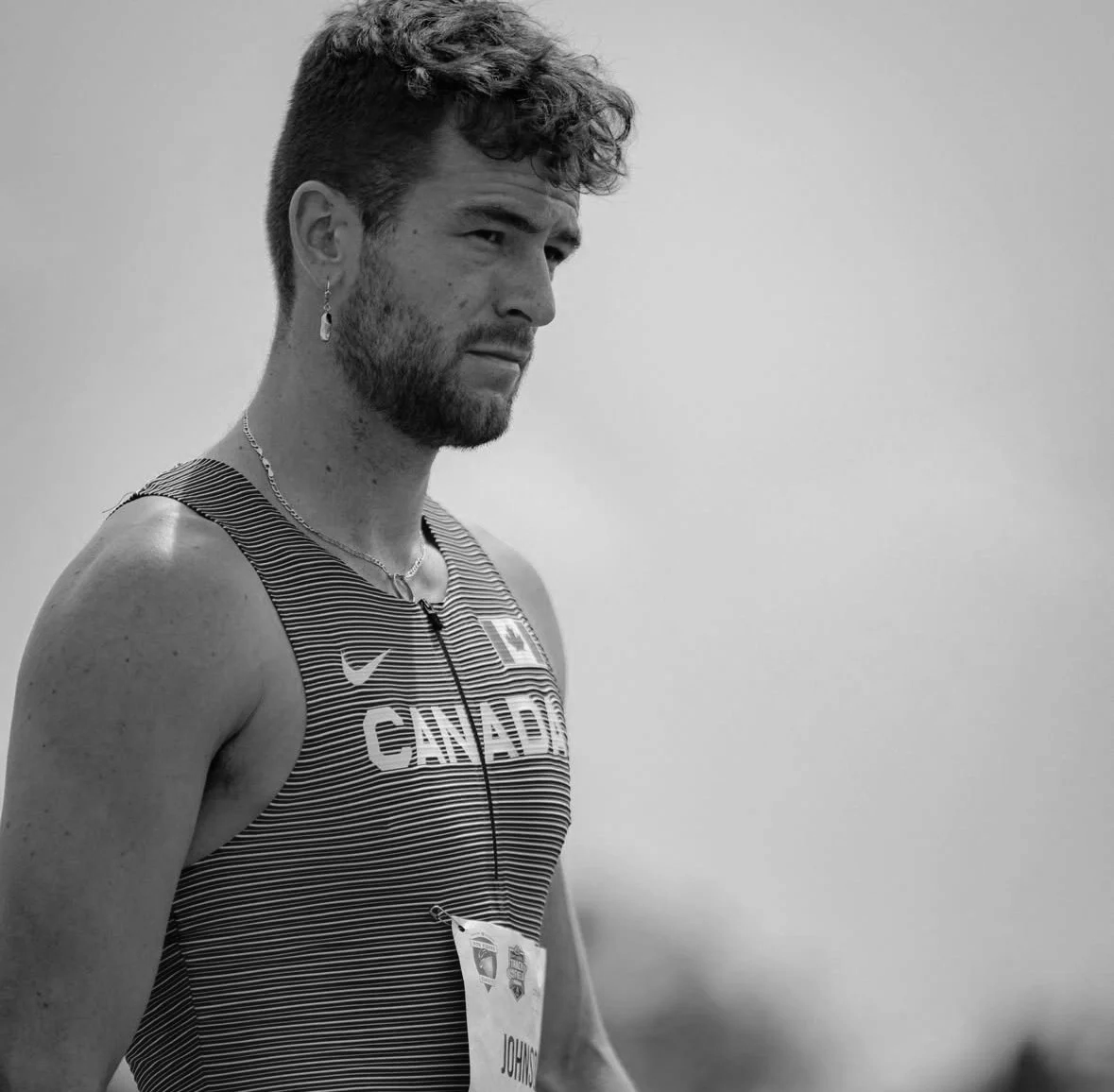 Black and white photo of a male athlete wearing a striped sports top with 'Canada' written on it, earring, necklace, and a bib number, standing outdoors.