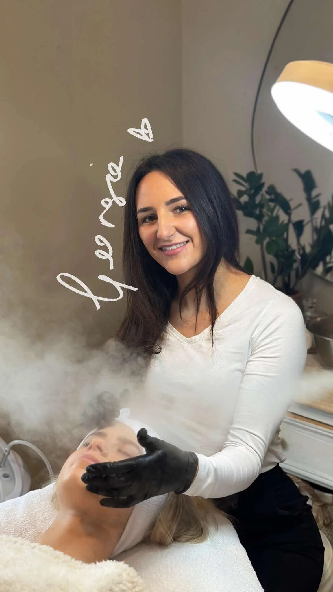 A woman with long black hair wearing a white long-sleeve shirt smiling at the camera while using a black gloved hand to perform a facial treatment on a woman lying down with closed eyes in a spa or skincare setting. The phrase 'love yourself' is written in white text with a heart symbol next to the woman.