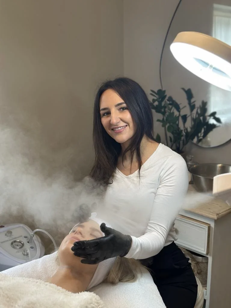 A beautician wearing gloves performing a facial treatment with steam on a client lying down in a spa or salon setting, with a lighted magnifying lamp and lush plant in the background.