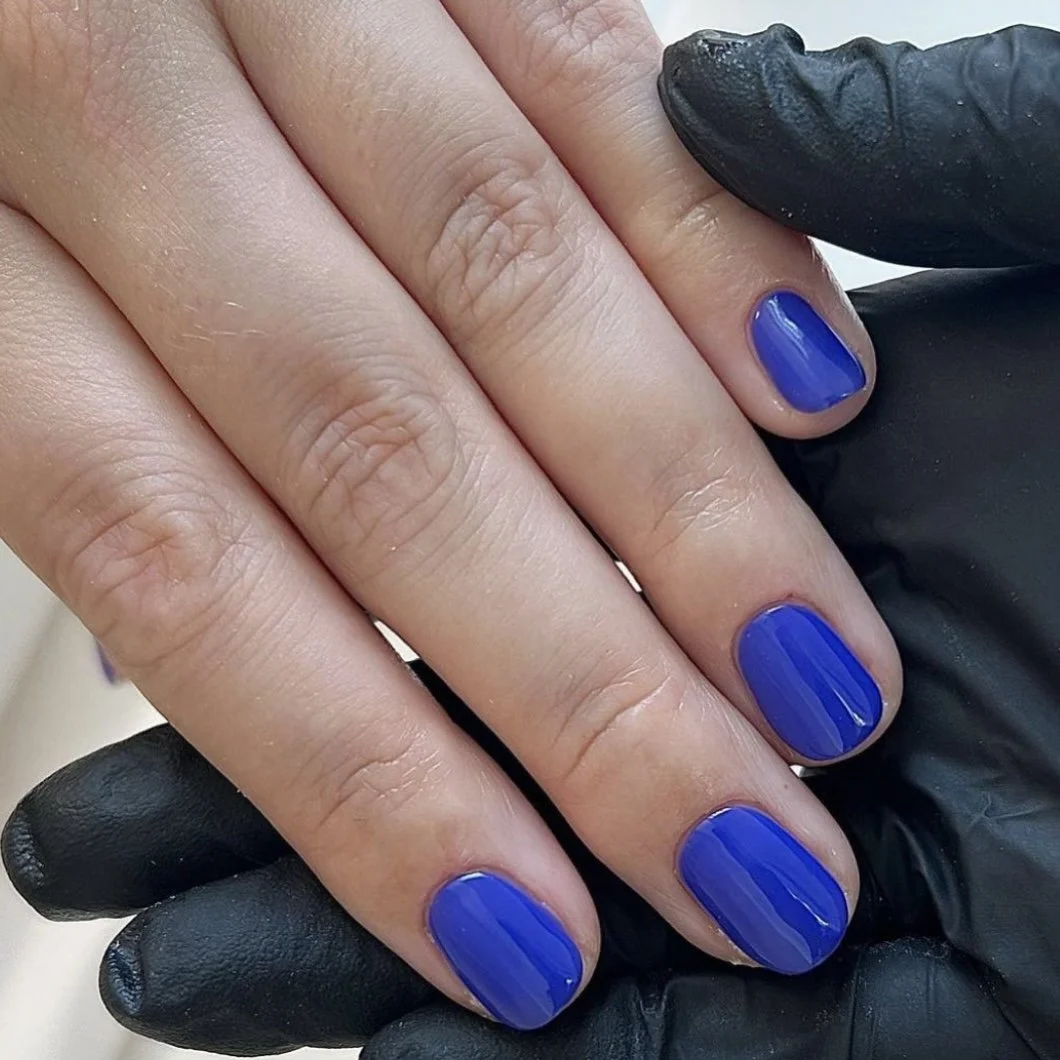 Close-up of a hand with short nails painted in vibrant blue color, with some skin detail visible, and a black glove partially covering the hand.