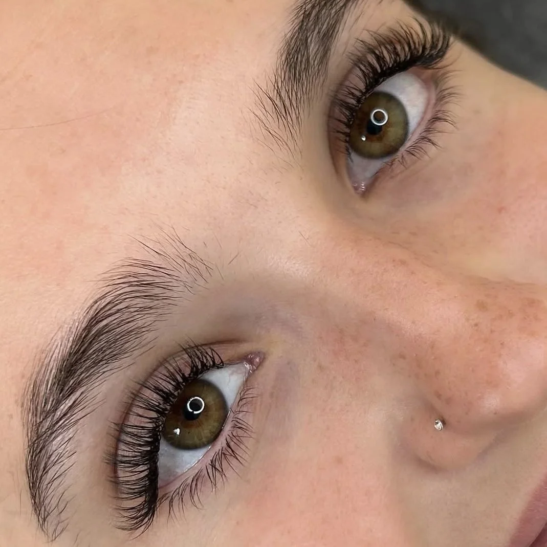 Close-up of person's brown eyes, with long eyelashes and a small nose piercing. The skin shows natural freckles.