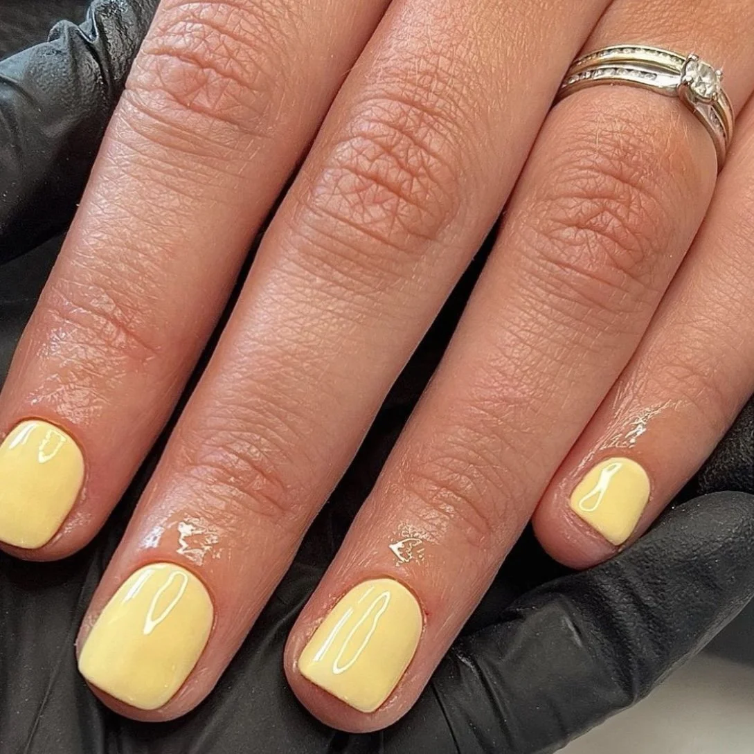 Close-up of a hand with yellow painted nails, a silver ring on the ring finger, and a black glove partially visible underneath.
