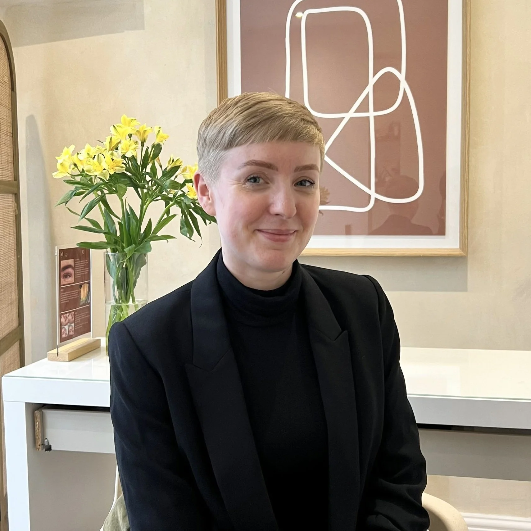 Person sitting in a modern room with abstract wall art, a vase of yellow flowers, and wearing a black blazer and turtleneck.