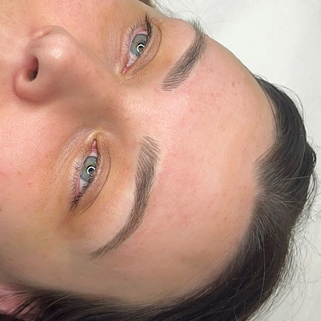 Close-up of a woman's face with freshly groomed eyebrows and light makeup, lying down on her side.