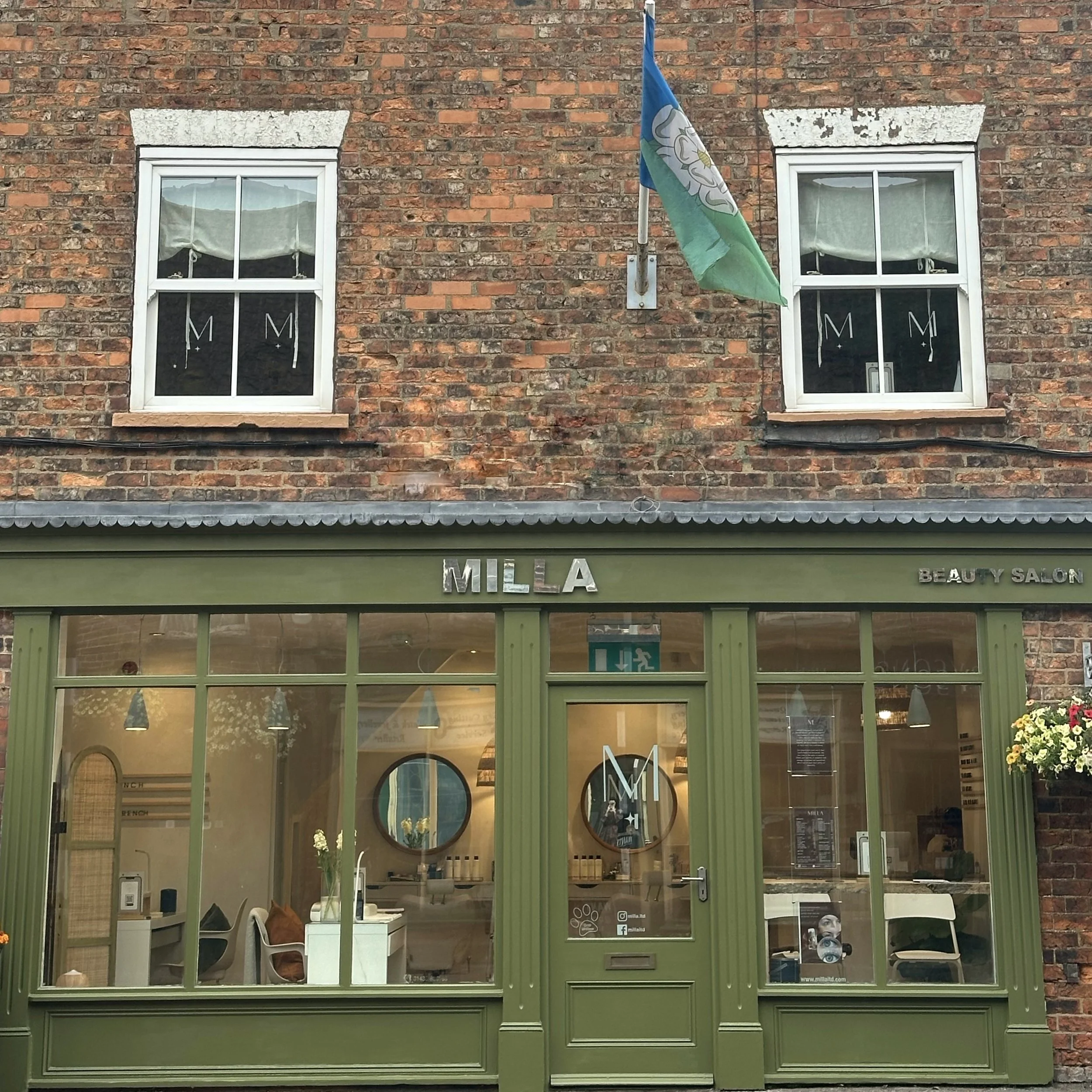 Green storefront with the sign 'MILLA' and 'Beauty Salon' on the right side, two second-story windows with white frames, and a flag flying above the entrance.
