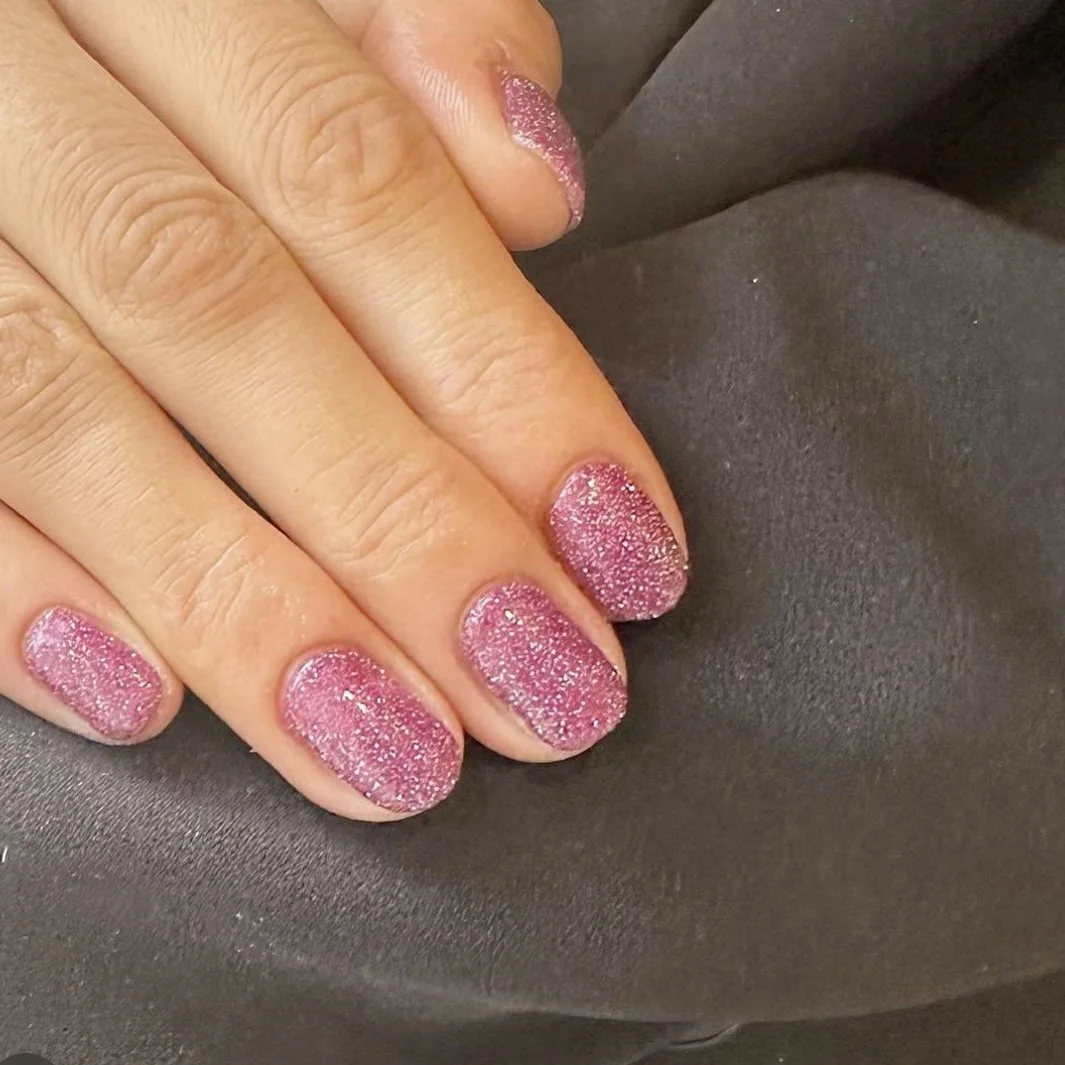 Close-up of a hand with nails painted pink glittery nail polish, resting on a dark fabric surface.
