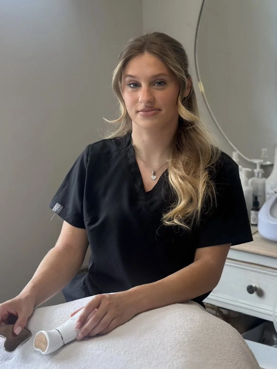 A young woman with long wavy blonde hair, wearing a black medical scrub top, sitting at a table with a handheld device in her hand, in a medical or spa setting.
