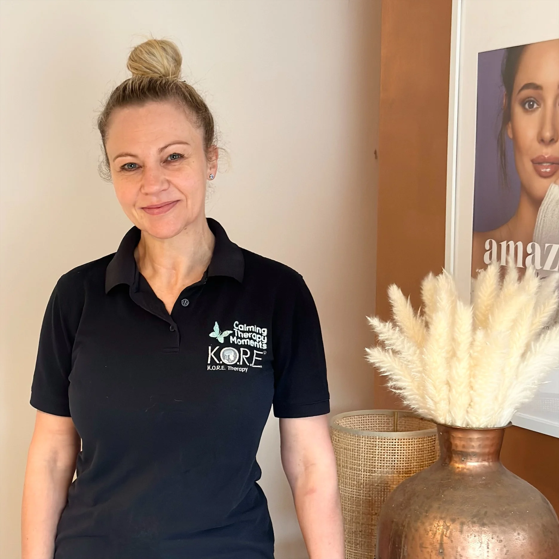 A woman wearing a black polo shirt with the logo 'Calming Therapy Moments KORE Therapy' stands smiling. She has blonde hair tied in a bun. Beside her is a large copper vase with beige pampas grass, and a partially visible poster in the background.