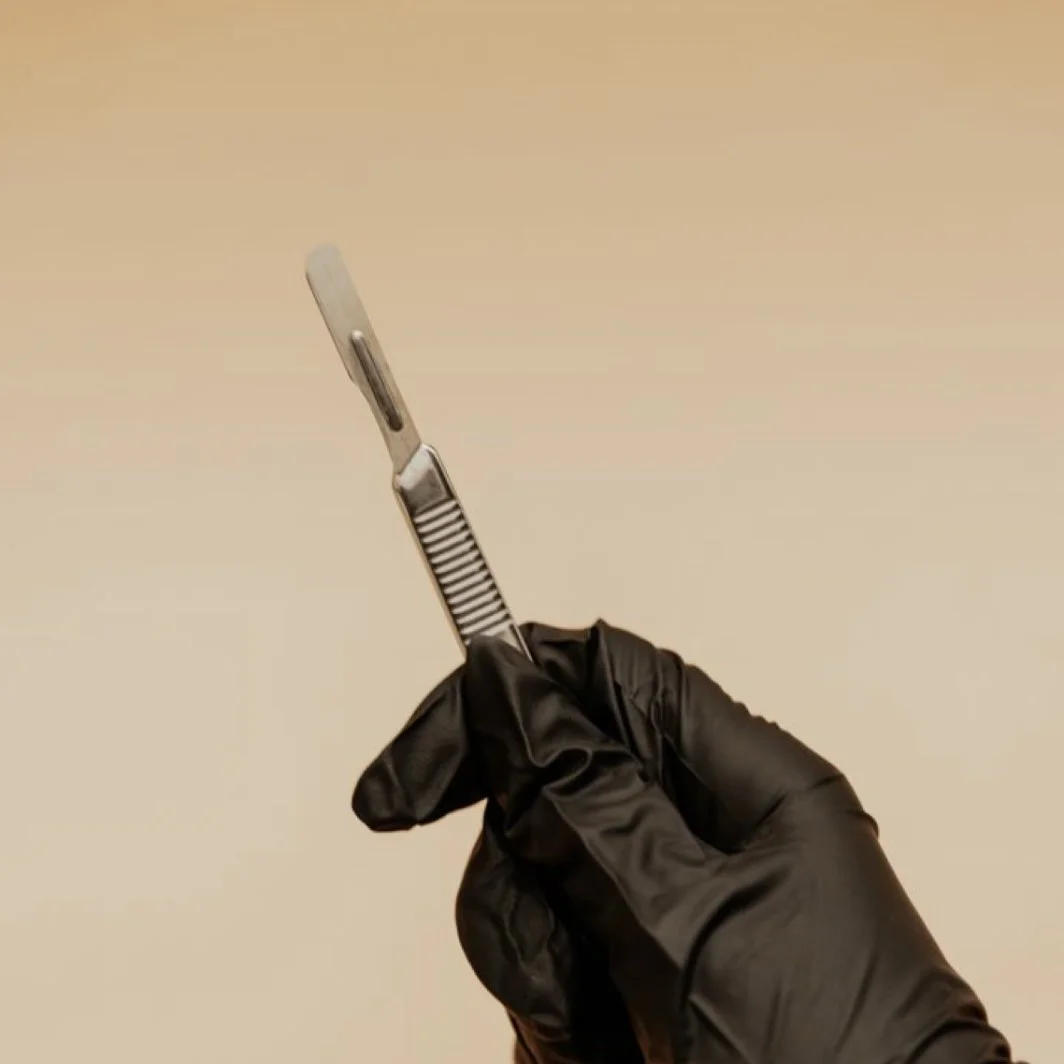A person wearing black gloves holding a scalpel against a beige background.