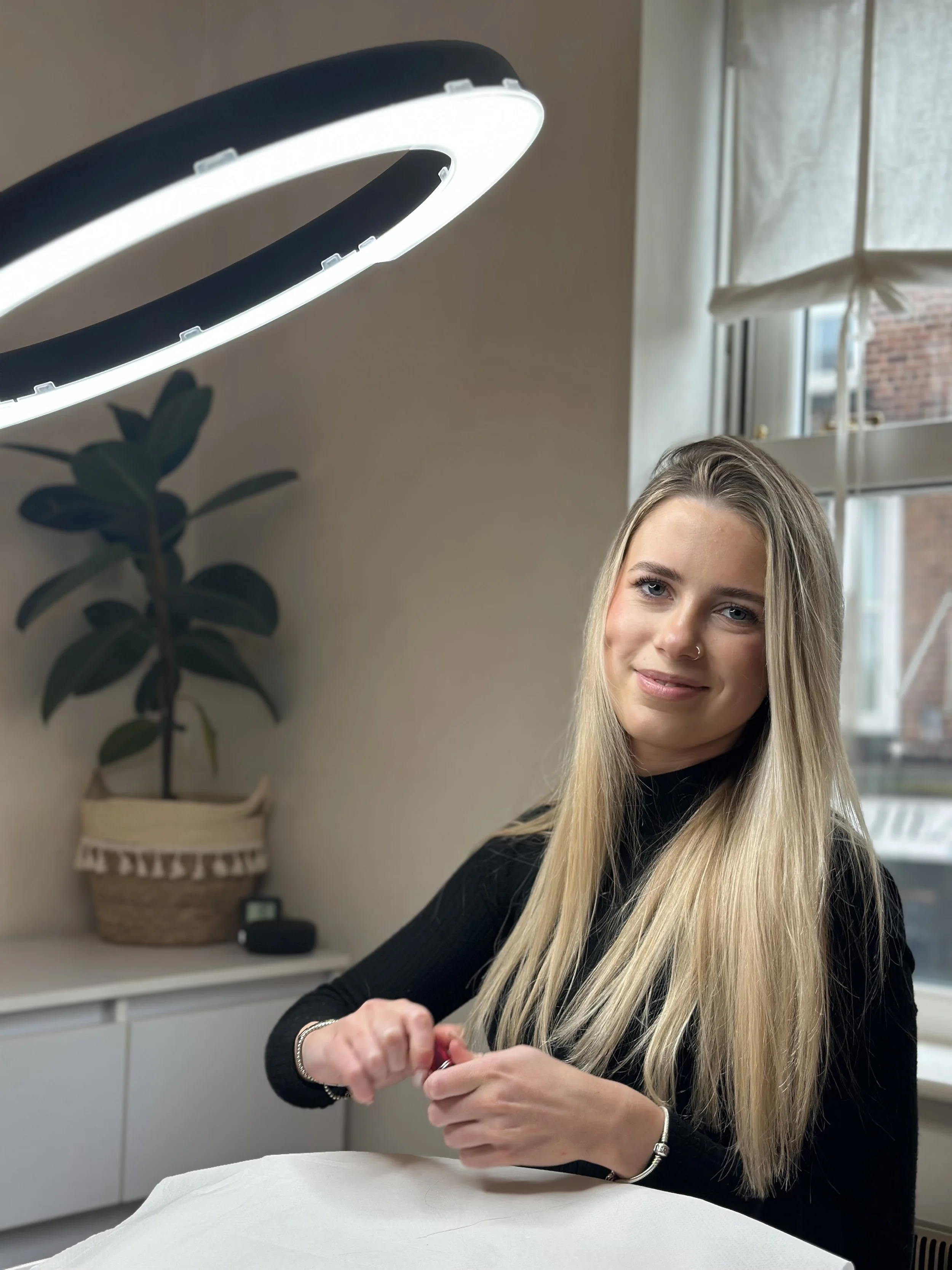 A young woman with long blonde hair sitting indoors near a window, smiling at the camera, under a circular LED ring light, with a plant and a basket behind her.