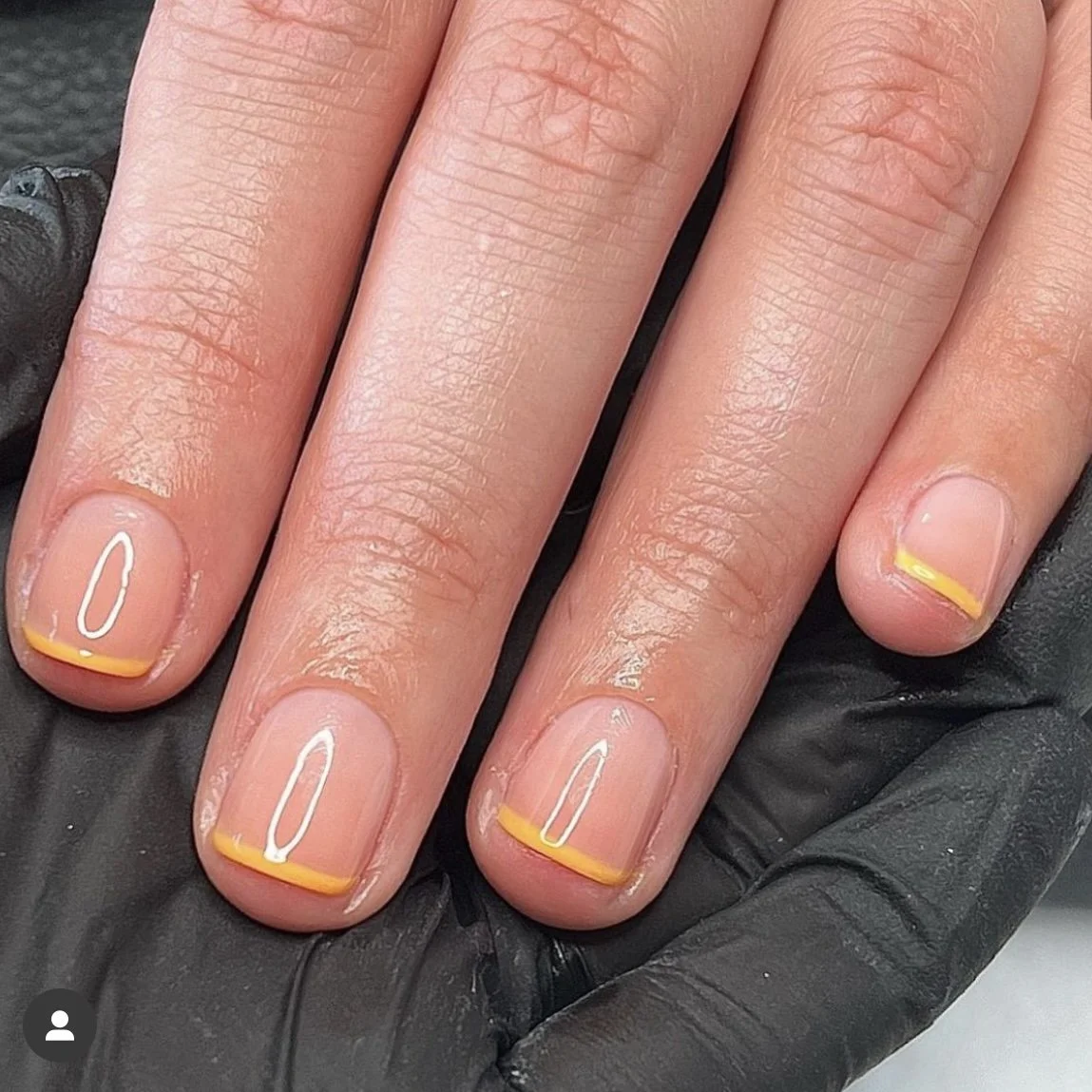 Close-up of a hand with neatly manicured nails. The nails have a clear base with a small yellow-orange border at the tip. The hand is resting on a black glove.