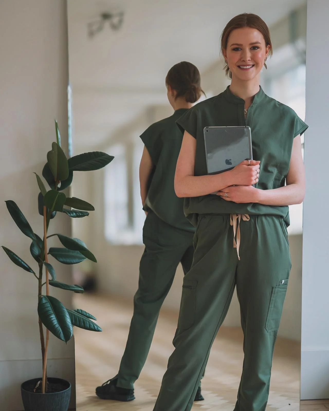 Woman in green outfit holding a tablet, standing in front of a mirror next to a potted plant.