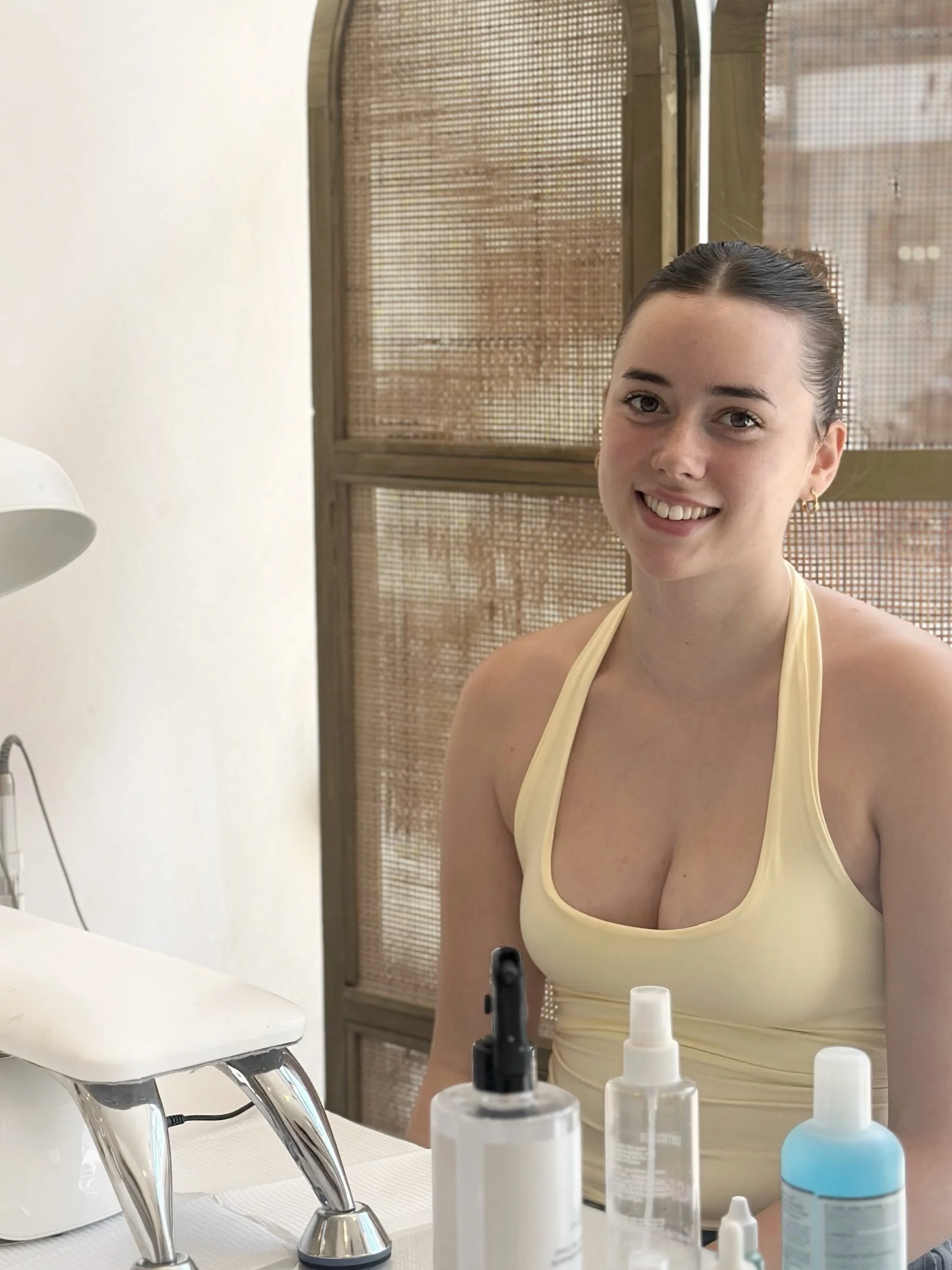 A woman with dark hair tied back, wearing a yellow tank top, sitting at a table with skincare products in bottles and sprays, smiling at the camera.