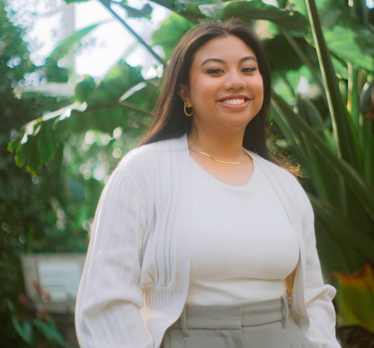 A young woman with long dark hair smiling, wearing a white top, jewelry, and standing outdoors surrounded by green plants.