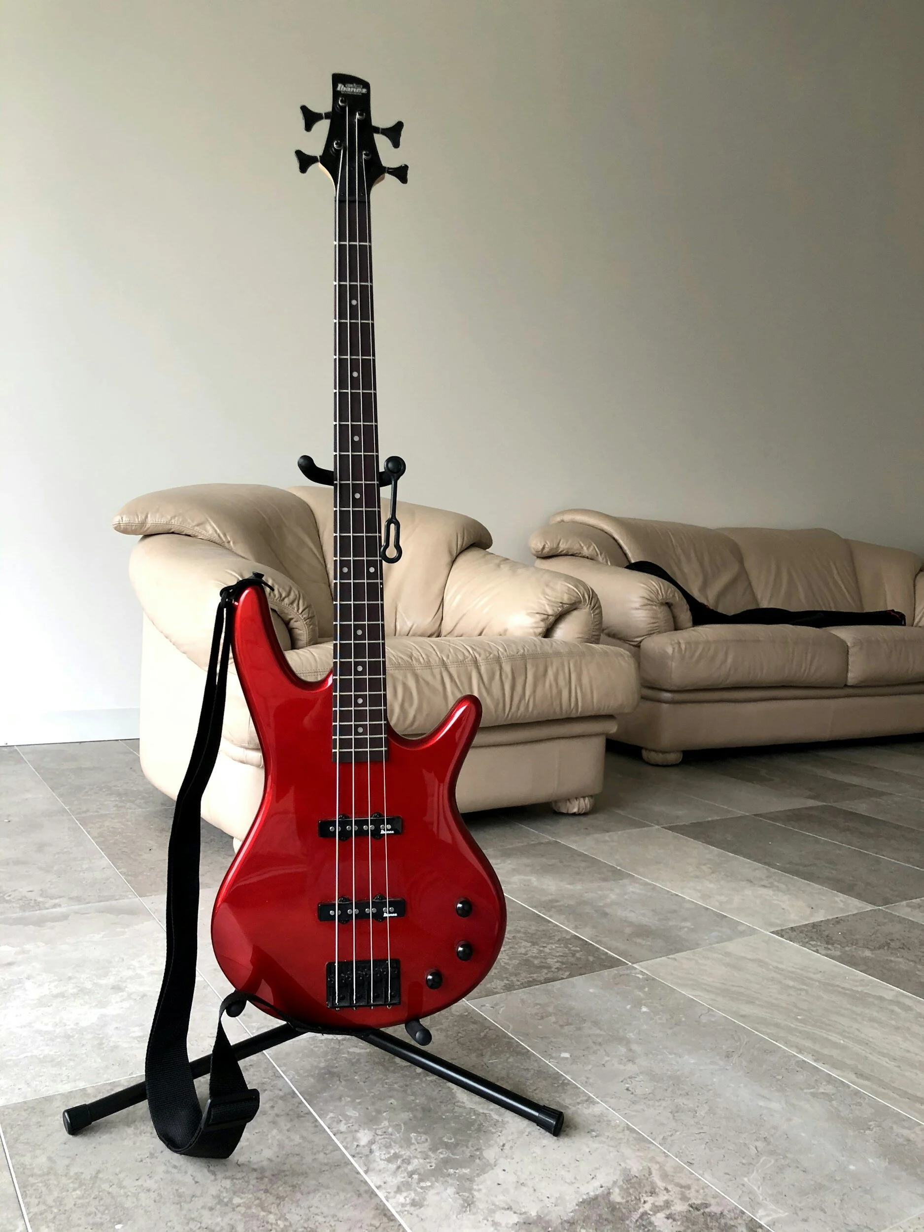 A red electric bass guitar with a strap on a stand in a living room with beige couches and a tiled floor.