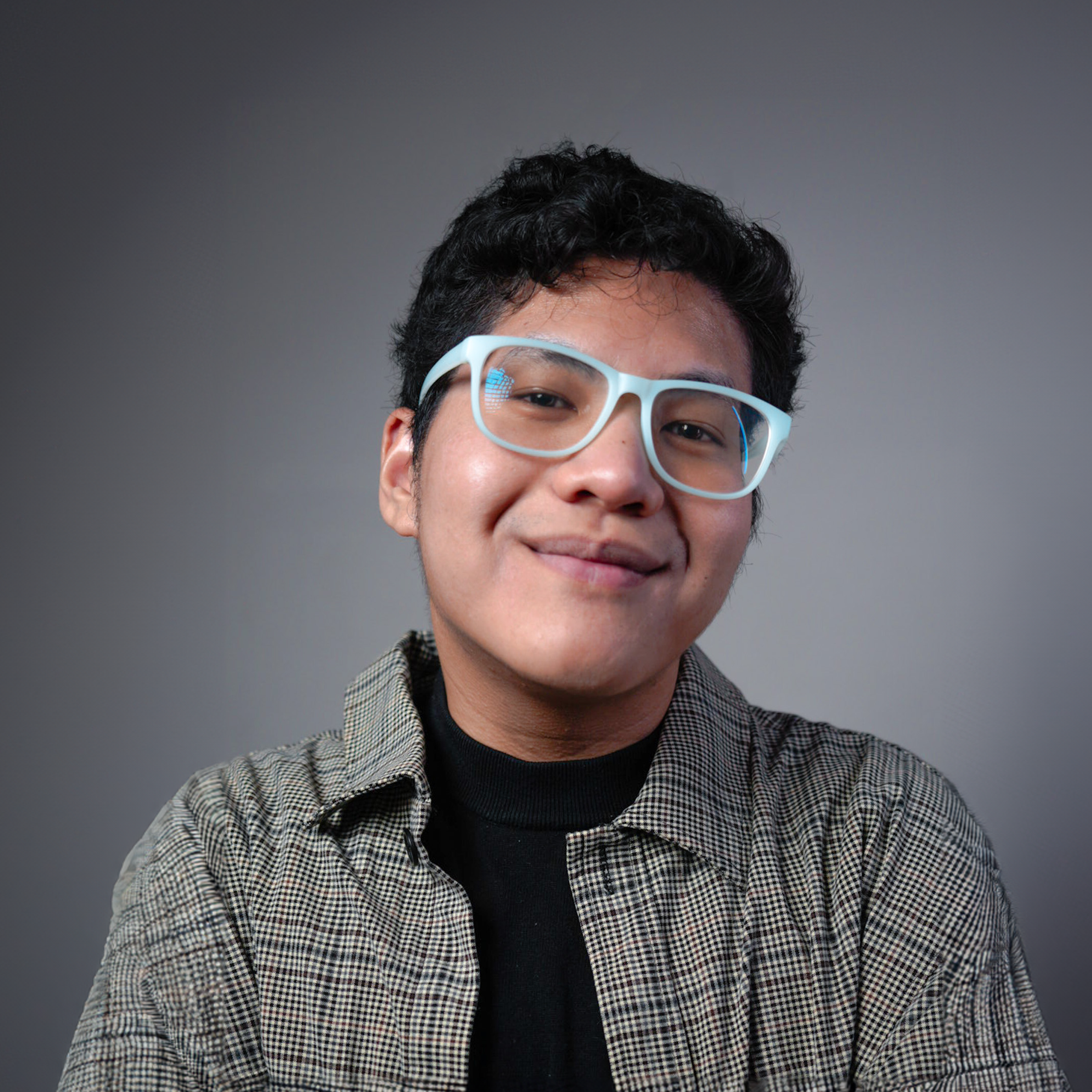 Young man wearing white glasses and a checkered shirt, smiling against a plain gray background.