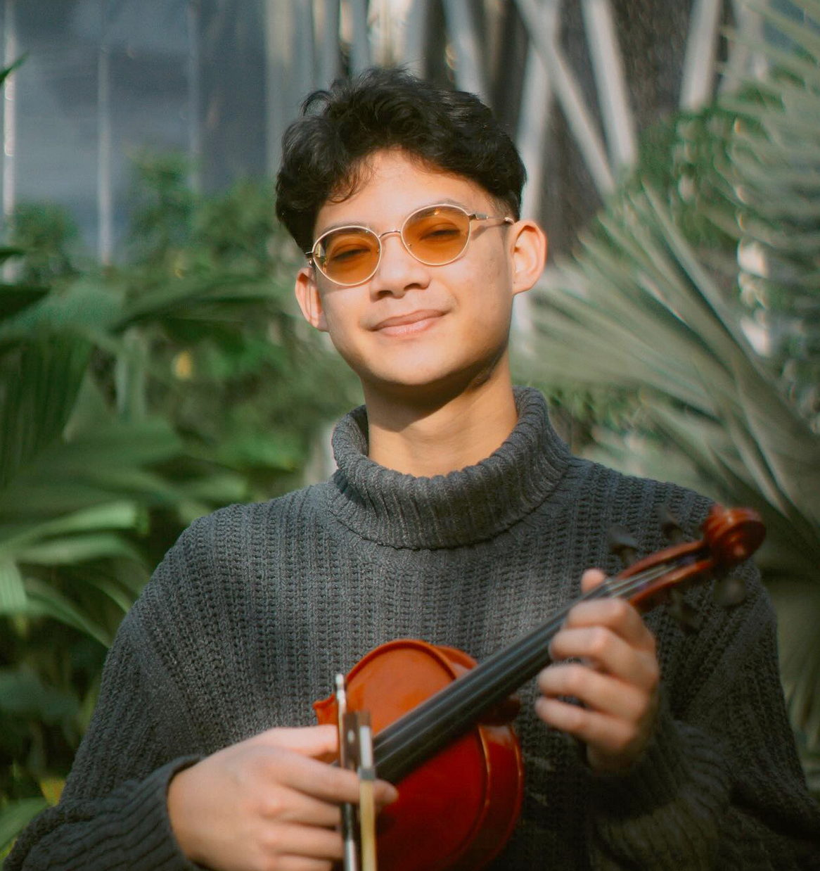 Young man wearing sunglasses and a gray turtleneck sweater, smiling, holding a violin in a lush green conservatory or greenhouse.