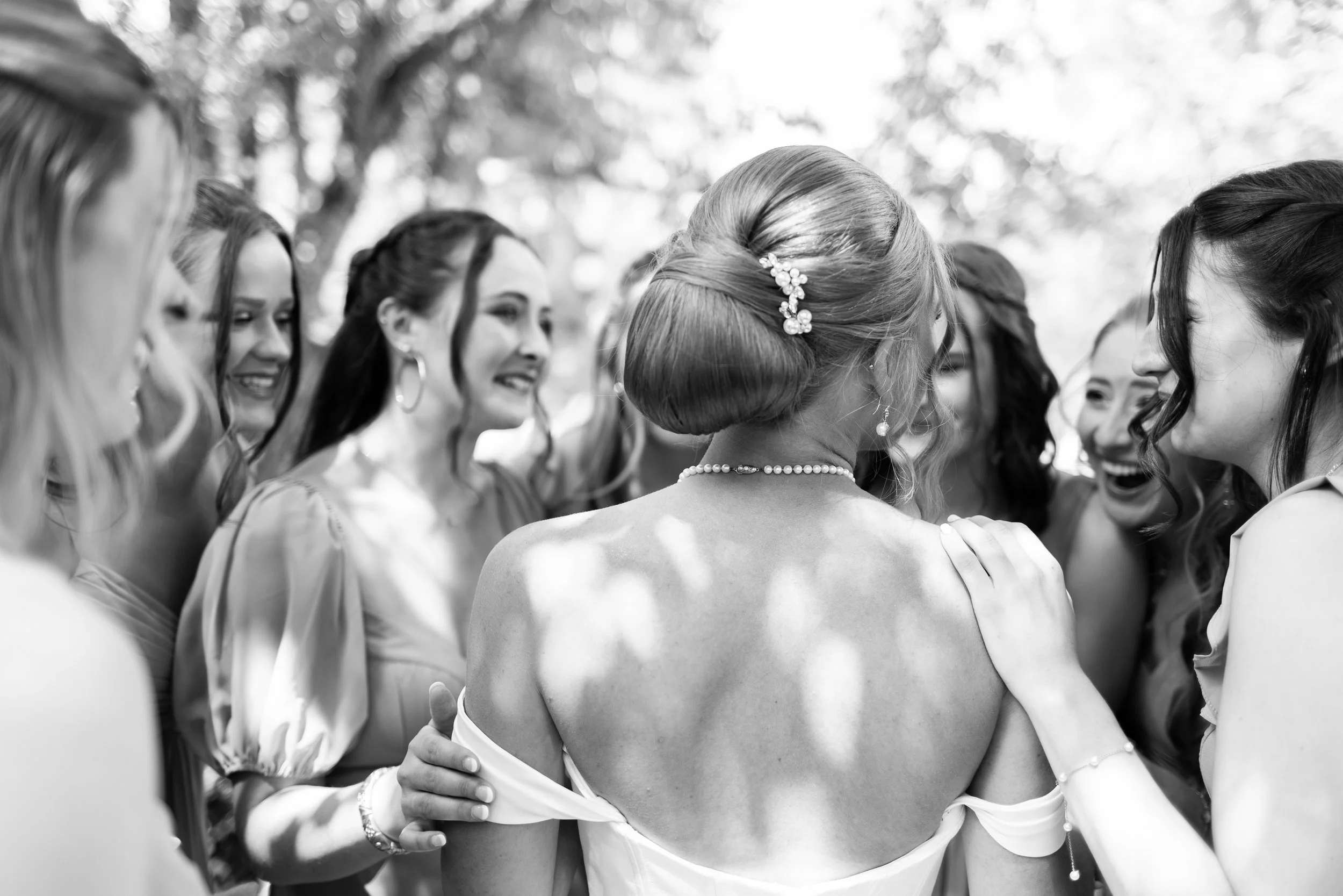 Black and white photo of several women gathered closely, with one woman in the center with her back to the camera. The women appear to be smiling and touching affectionately, suggesting a celebratory or intimate moment.