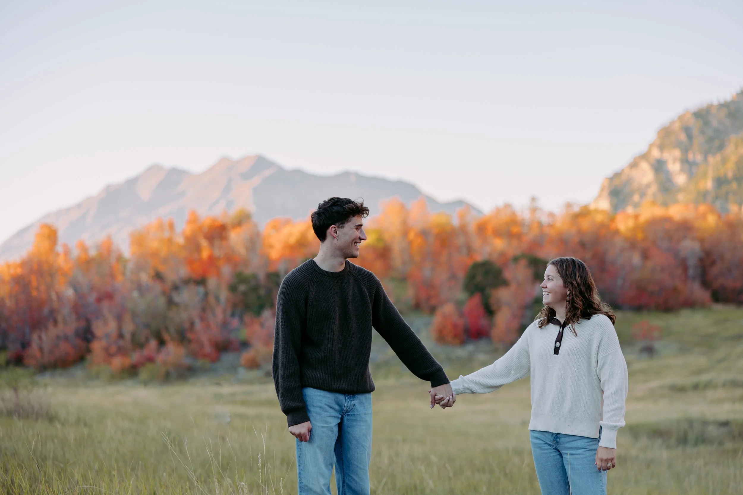 A young man and woman holding hands outdoors in a field, with autumn trees and mountains in the background.