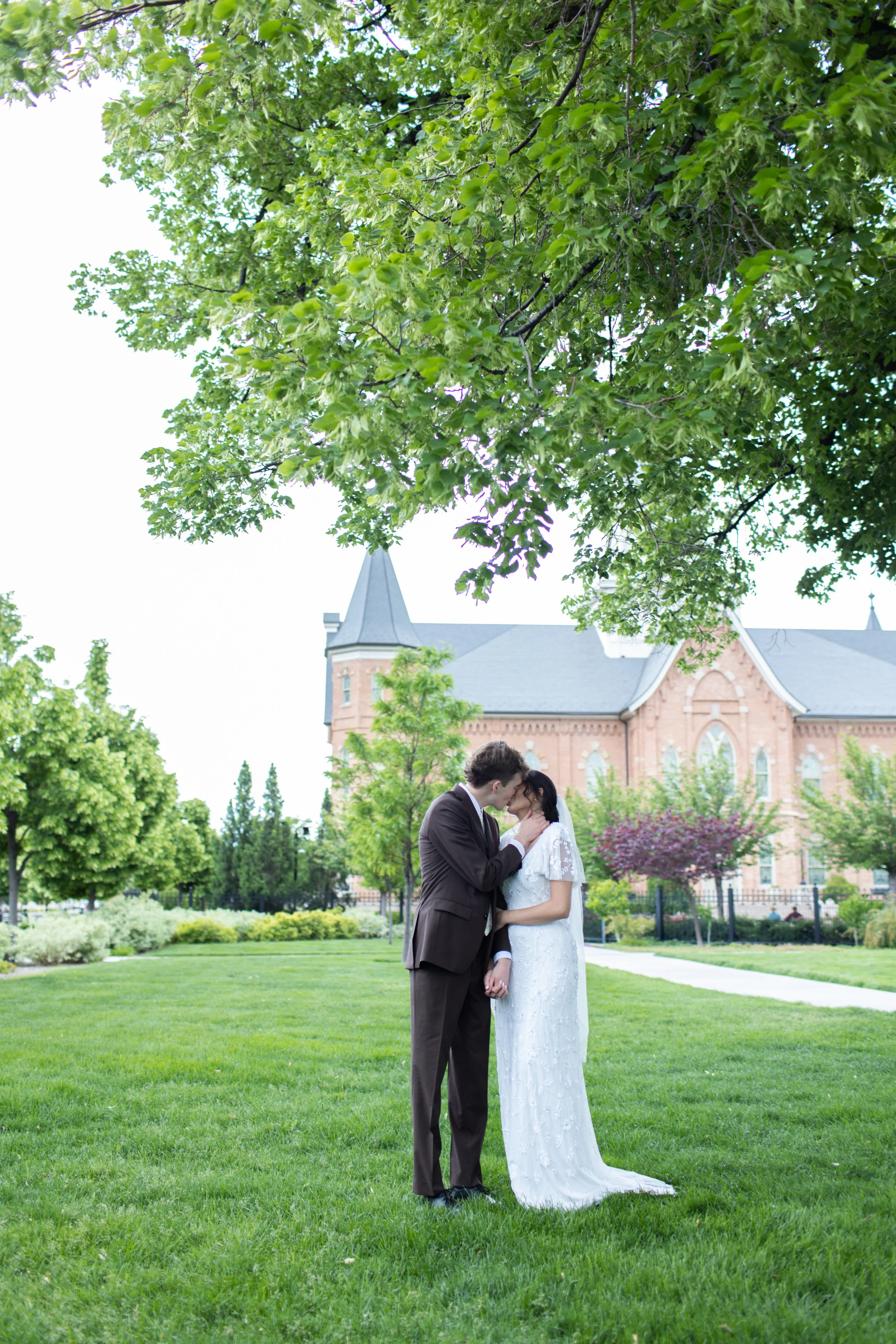 A couple dressed in wedding attire stands close together outdoors on a grassy lawn beneath a large leafy tree, sharing an intimate moment with a historic building in the background.