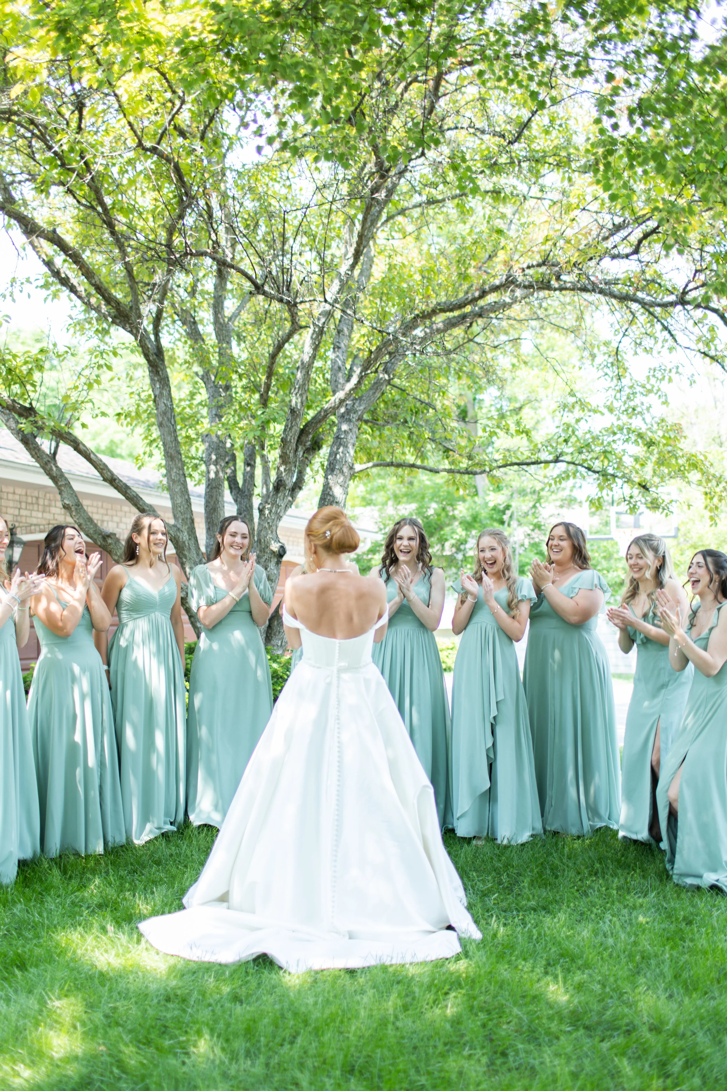 A bride in a white wedding dress standing outdoors with her back to the camera, surrounded by smiling bridesmaids dressed in matching mint green dresses, under a large tree with green leaves.