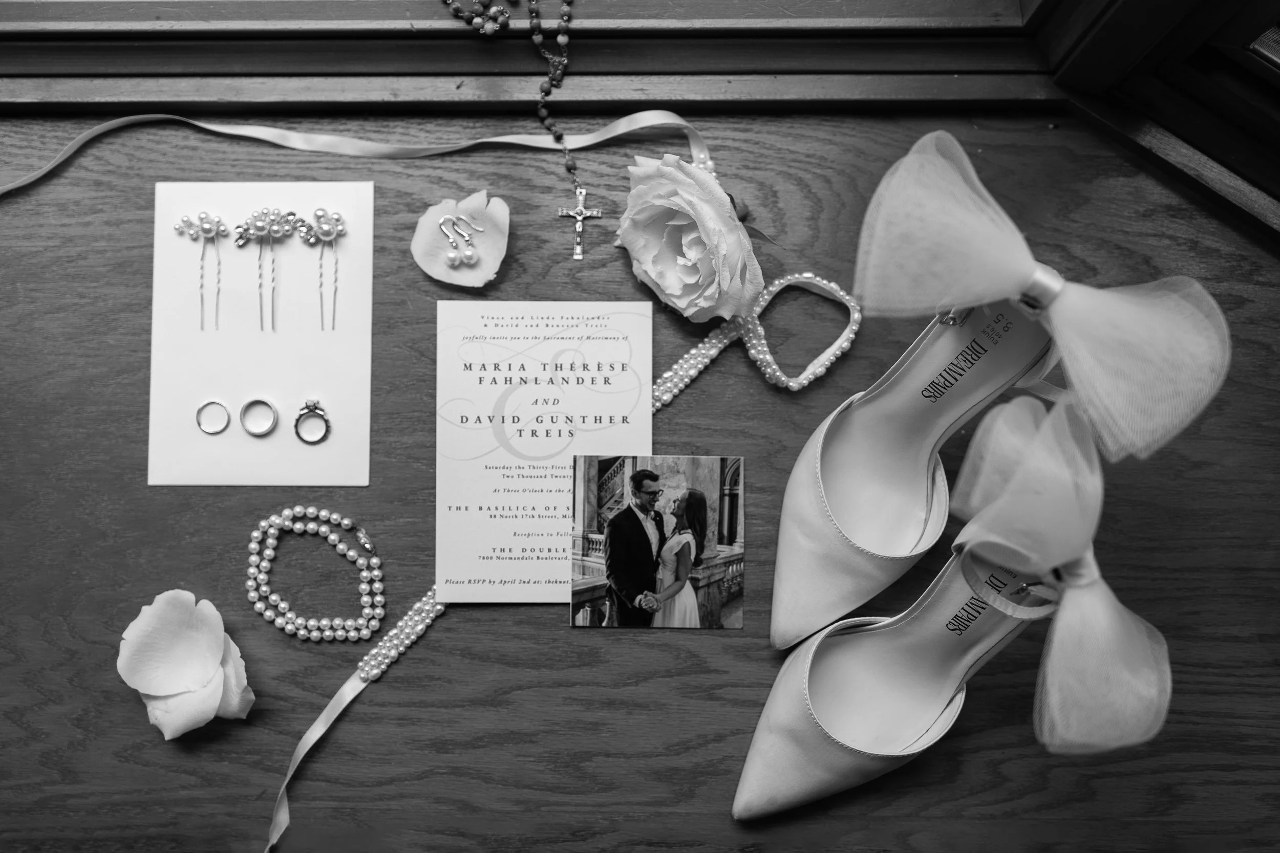 Black and white photo of wedding accessories and invitations on a wooden surface. Items include pearl earrings, a pearl necklace, a white rose, a pair of white high-heeled shoes, a wedding invitation, and other jewelry.