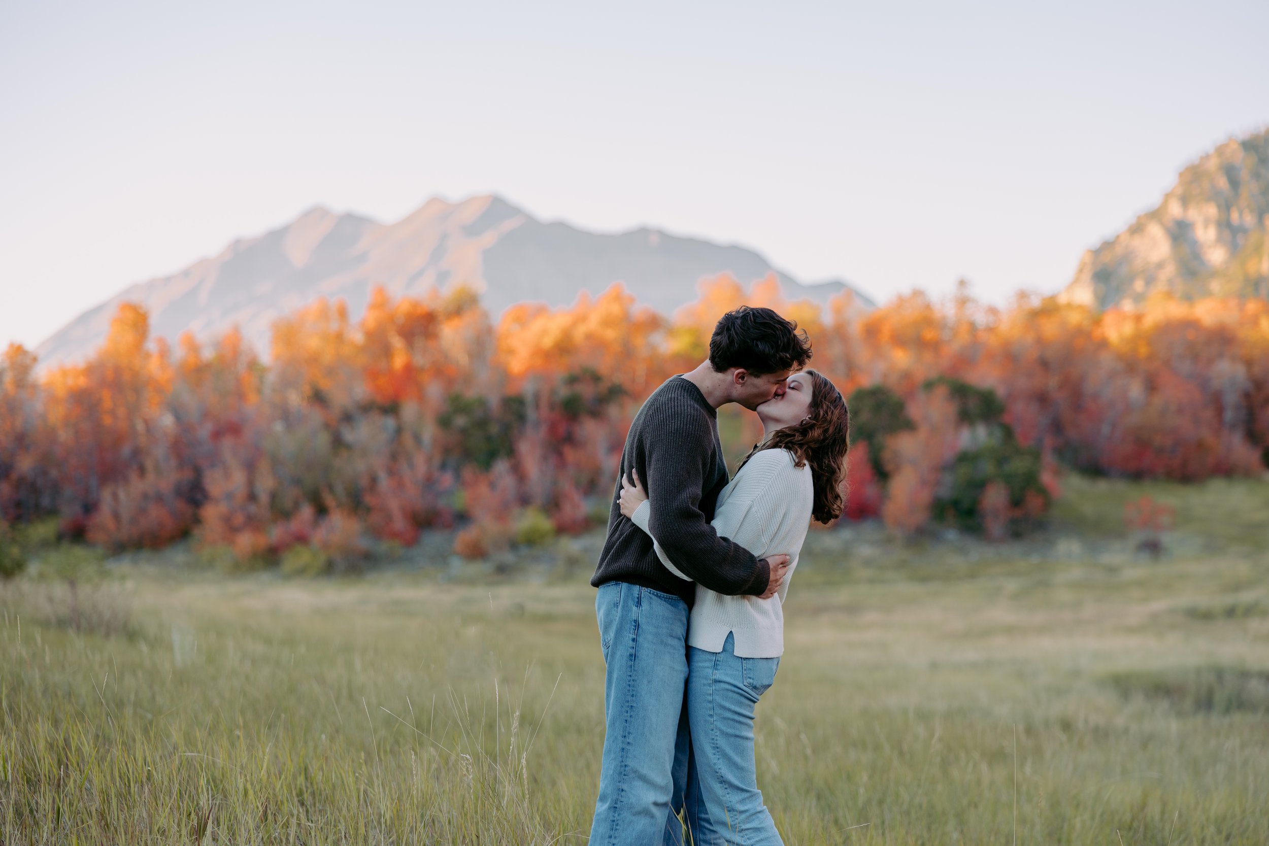 A couple sharing a kiss outdoors during autumn, with colorful fall trees and mountains in the background.