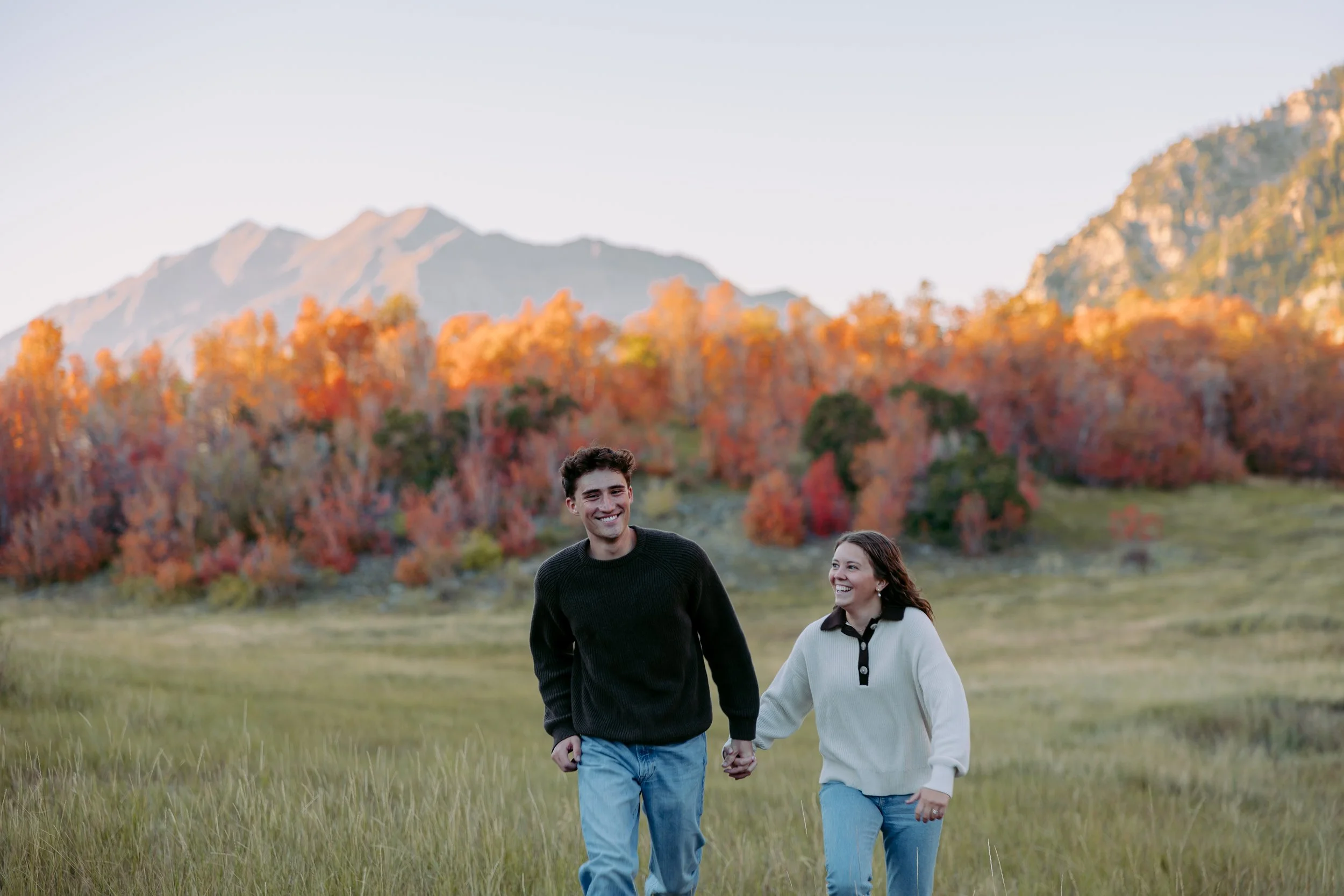 A young man and woman are walking hand in hand through a grassy field with colorful autumn trees and mountains in the background.