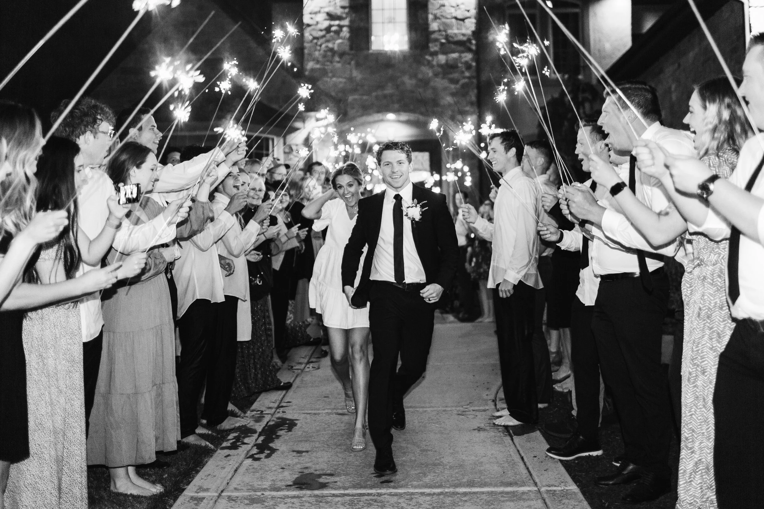 A black and white photo of a man and woman walking through a crowd holding sparklers during a celebration, possibly a wedding reception or party, on a city street at night.