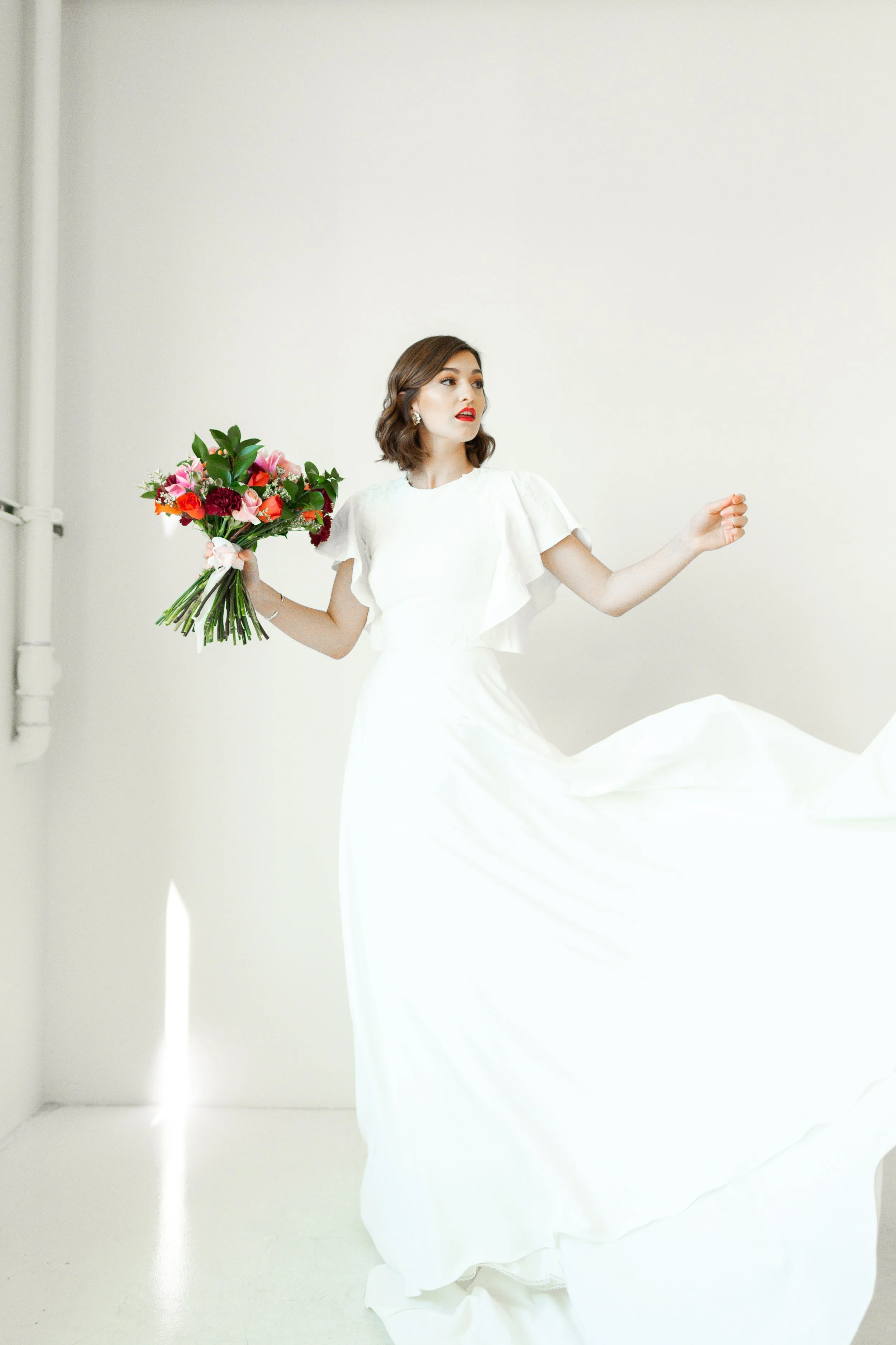 A woman in a white dress holding a bouquet of colorful flowers, standing against a plain, light-colored wall.
