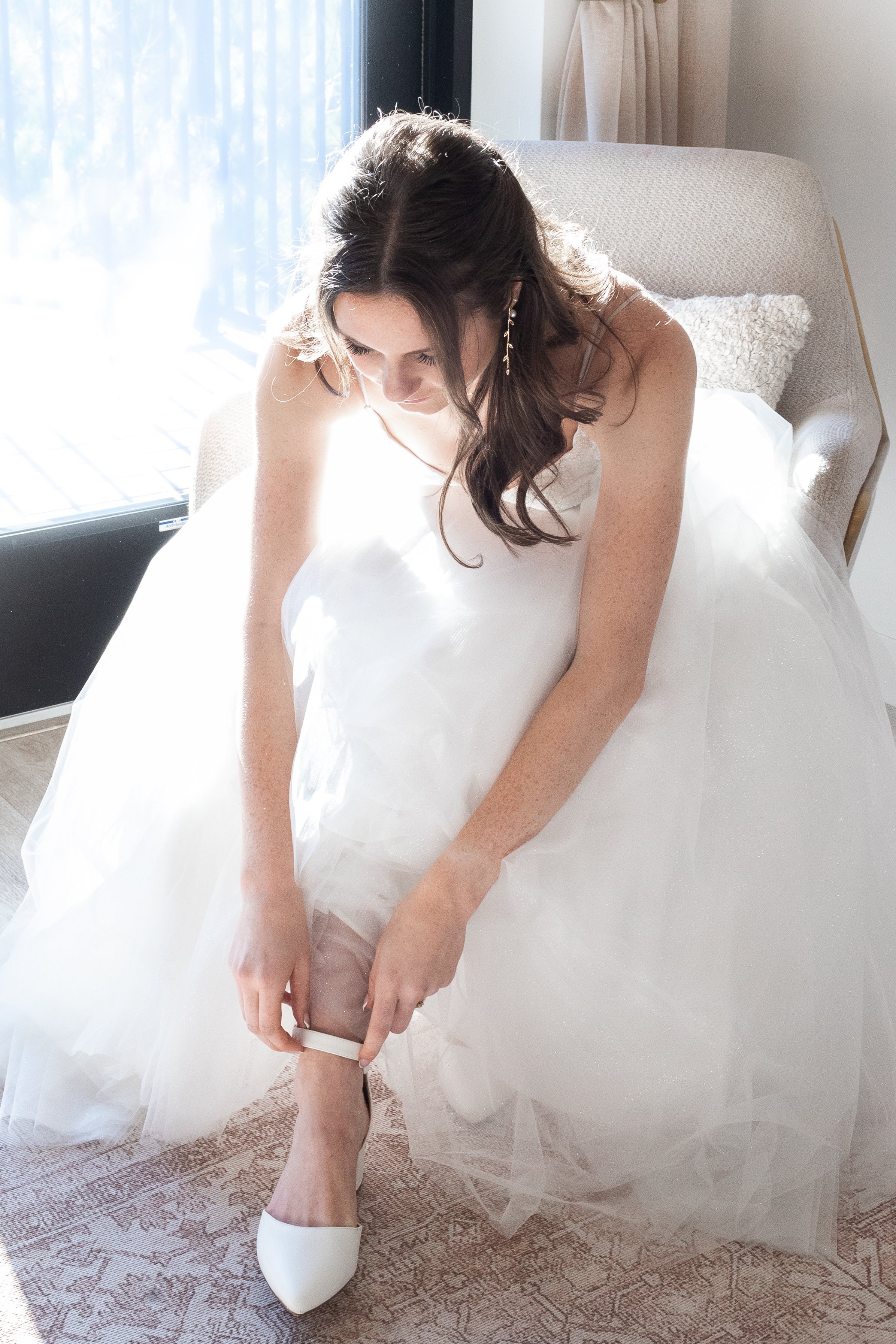 A woman in a wedding dress is sitting on a chair near a window with sunlight coming in. She is adjusting her white wedding shoe.