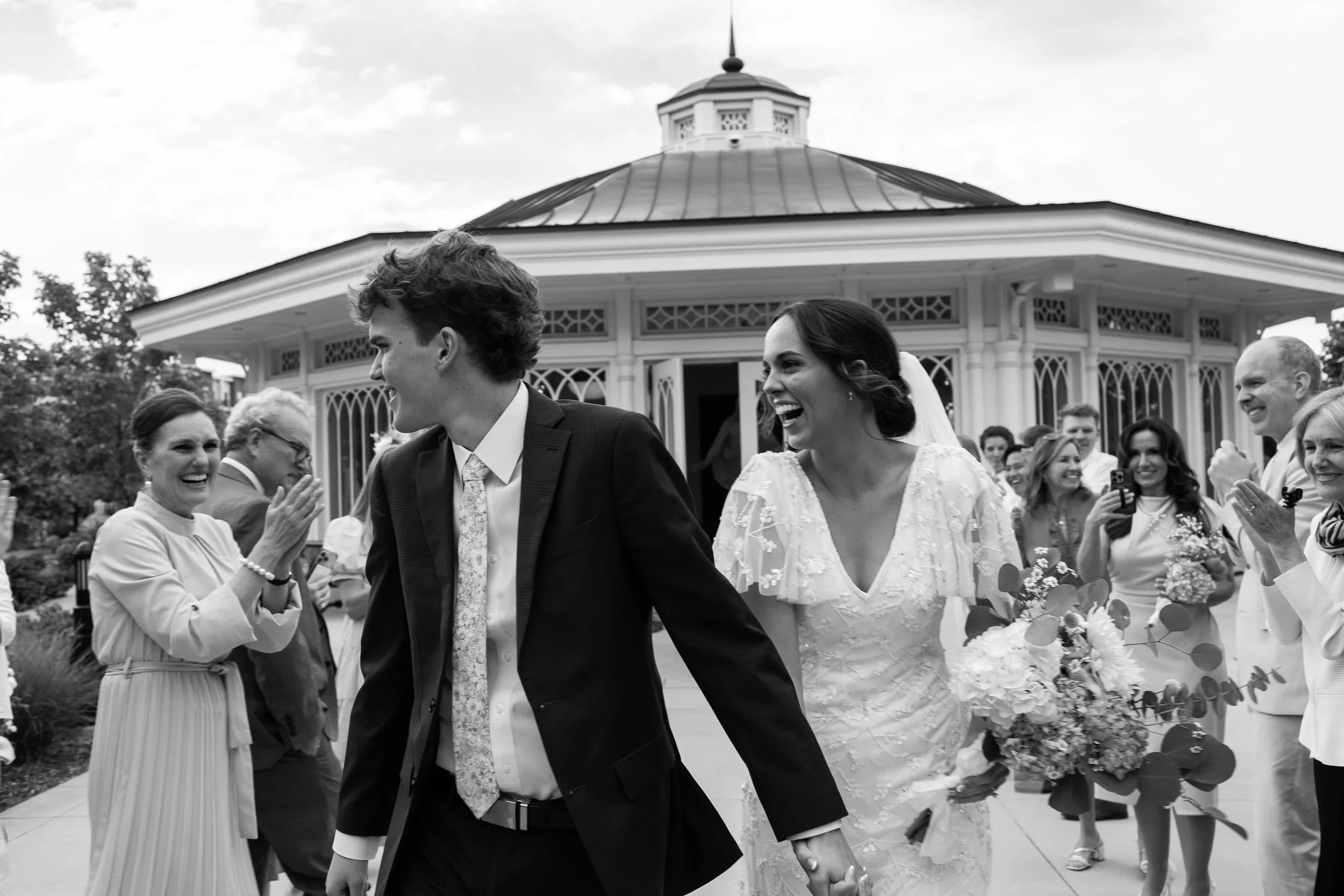 A black and white photo of a newlywed couple holding hands and smiling as they walk outdoors, surrounded by family and friends during a wedding celebration.