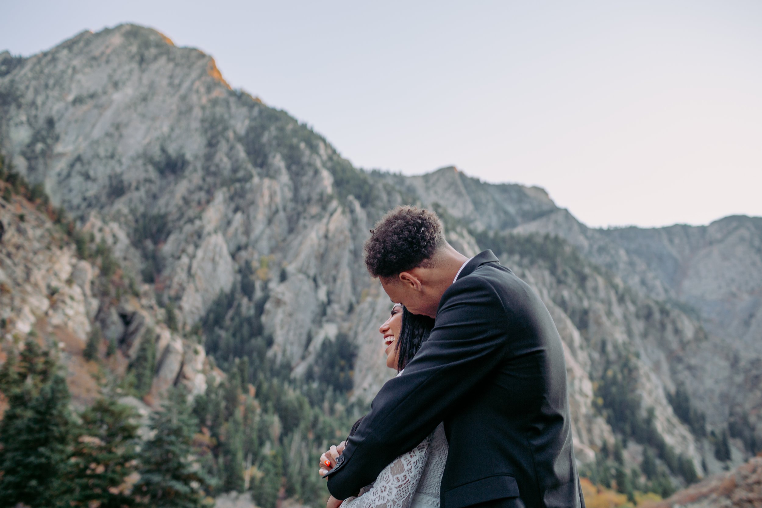 A couple embracing outdoors with mountains in the background, the man is leaning over the woman who is smiling.