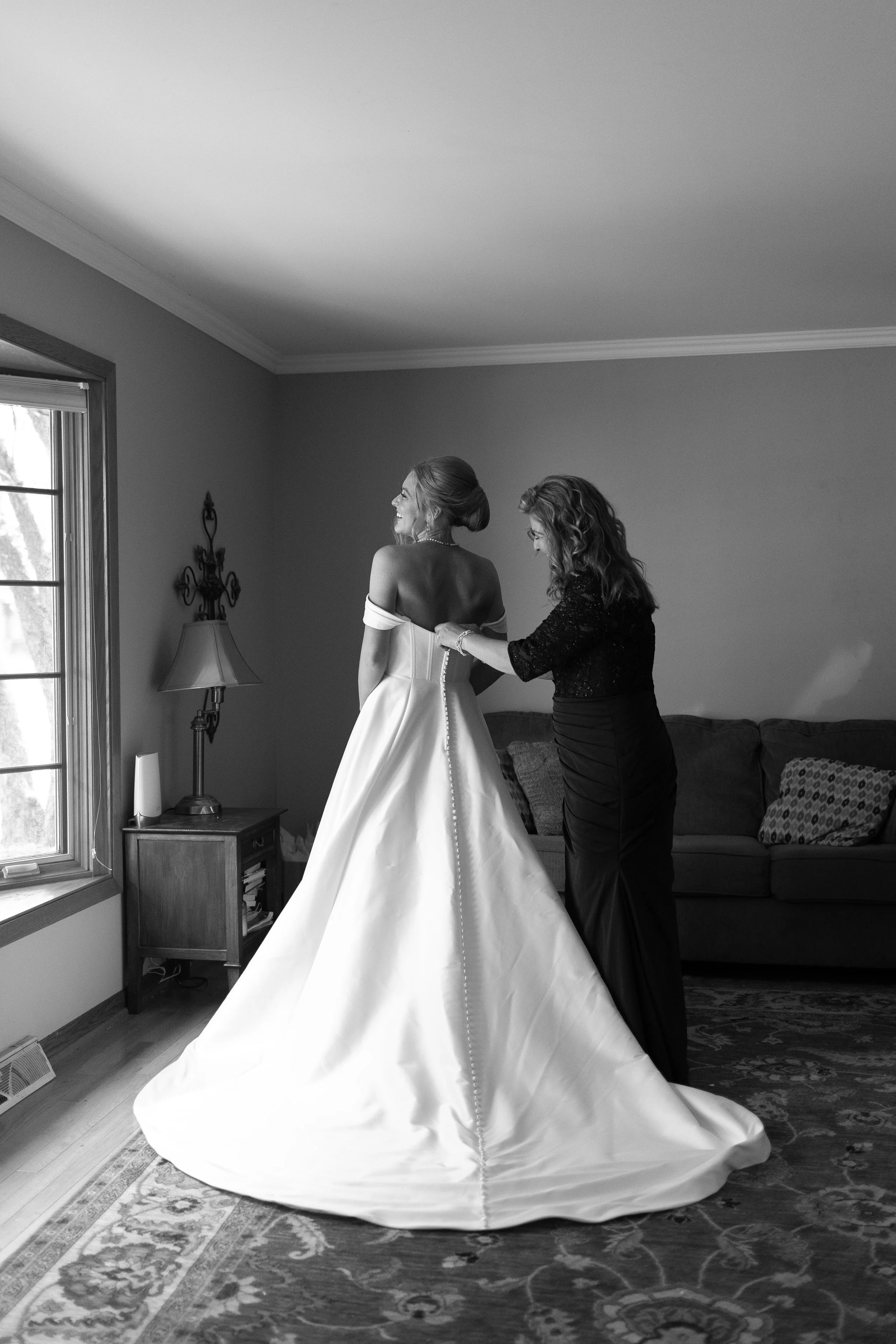 A bride standing in a room, helping her into her wedding dress with the assistance of another woman. The bride has her hair styled up and is smiling, while the other woman is adjusting the dress. There is a window with blinds, a small wooden table wi