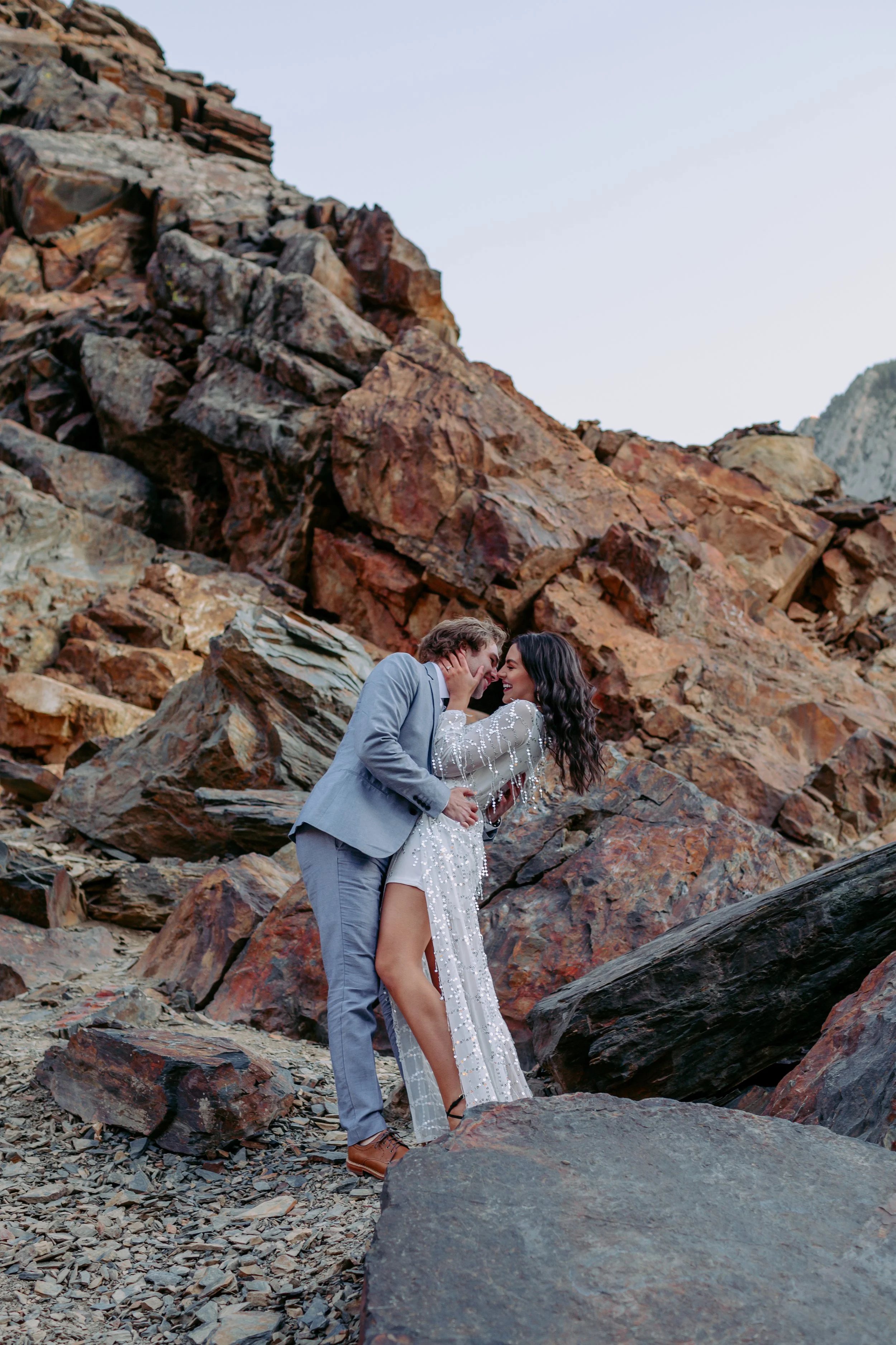 A couple dressed in wedding attire standing close and smiling on a rocky beach with large boulders and cliffs in the background.