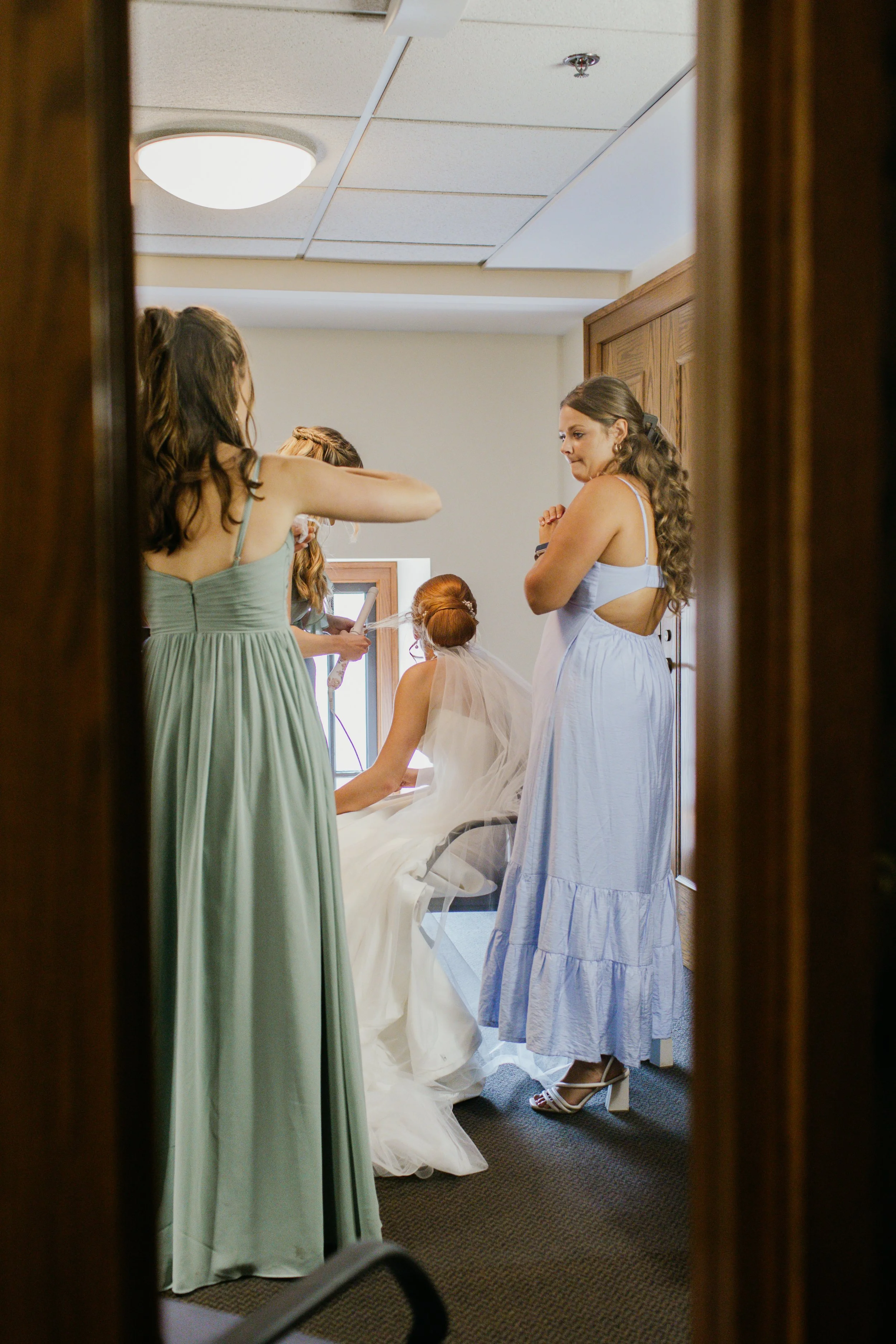 View of women in dresses preparing for a wedding, gathered around a bride seated on a chair, in a room with wood-paneled walls and a ceiling with lights and vents, seen through a doorway.