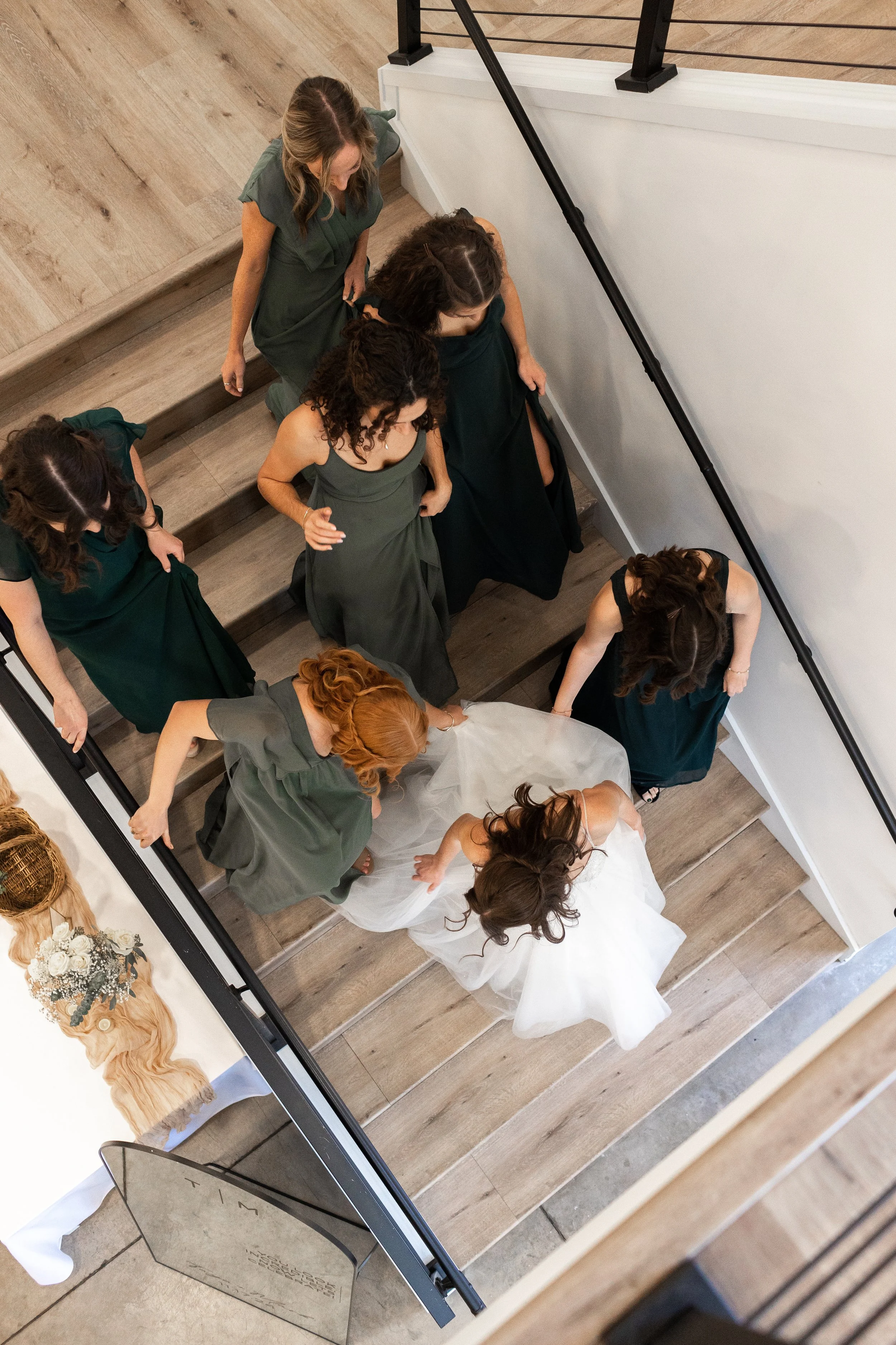 Multiple women dressed in formal attire, some in green dresses and others in darker gowns, descending a wooden staircase at a wedding or formal event.