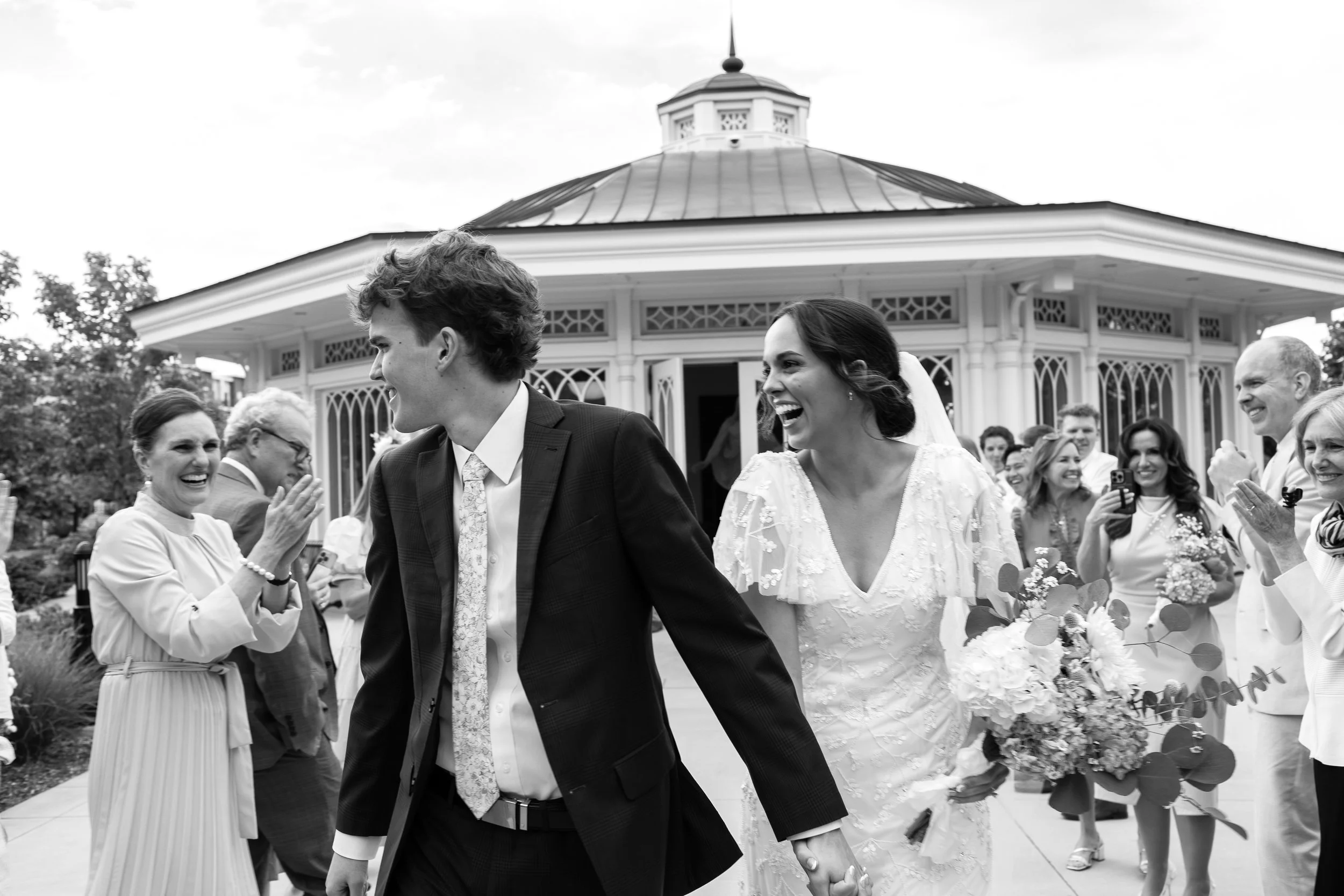 A black-and-white photo of a wedding celebration featuring a bride and groom walking outdoors, holding hands, surrounded by smiling guests, with a pavilion structure in the background.