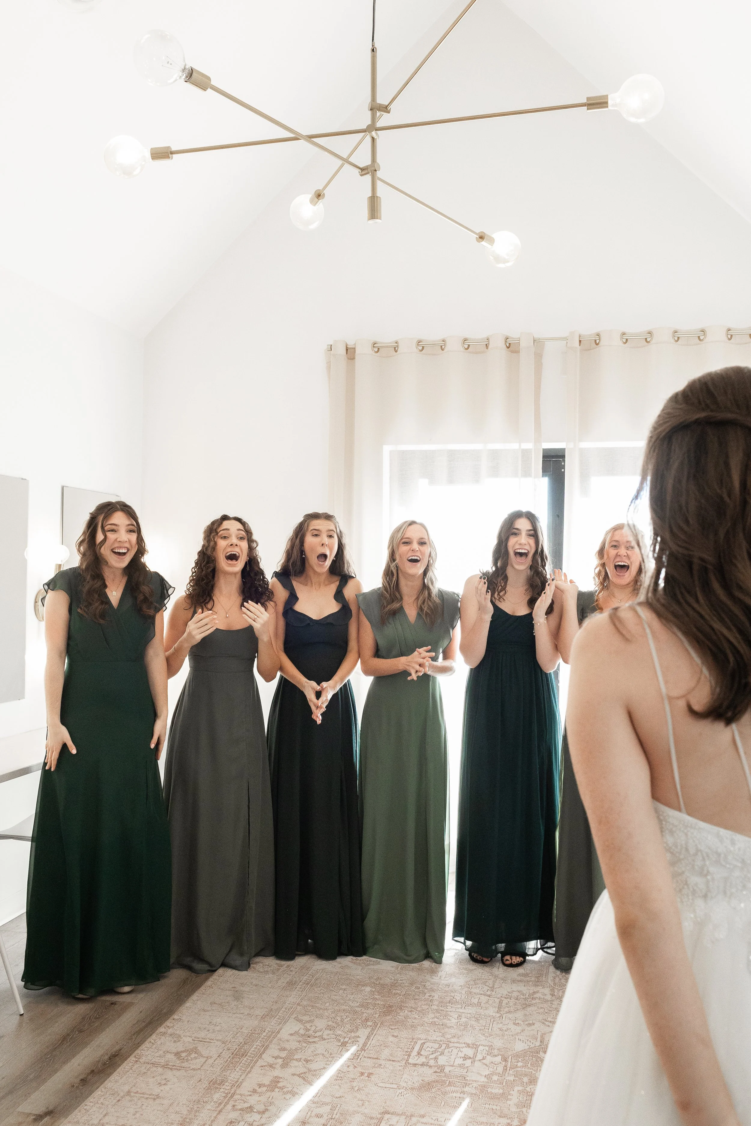 Group of women in formal dresses, standing indoors, smiling and laughing, with a white curtain and a modern ceiling light fixture in the background.