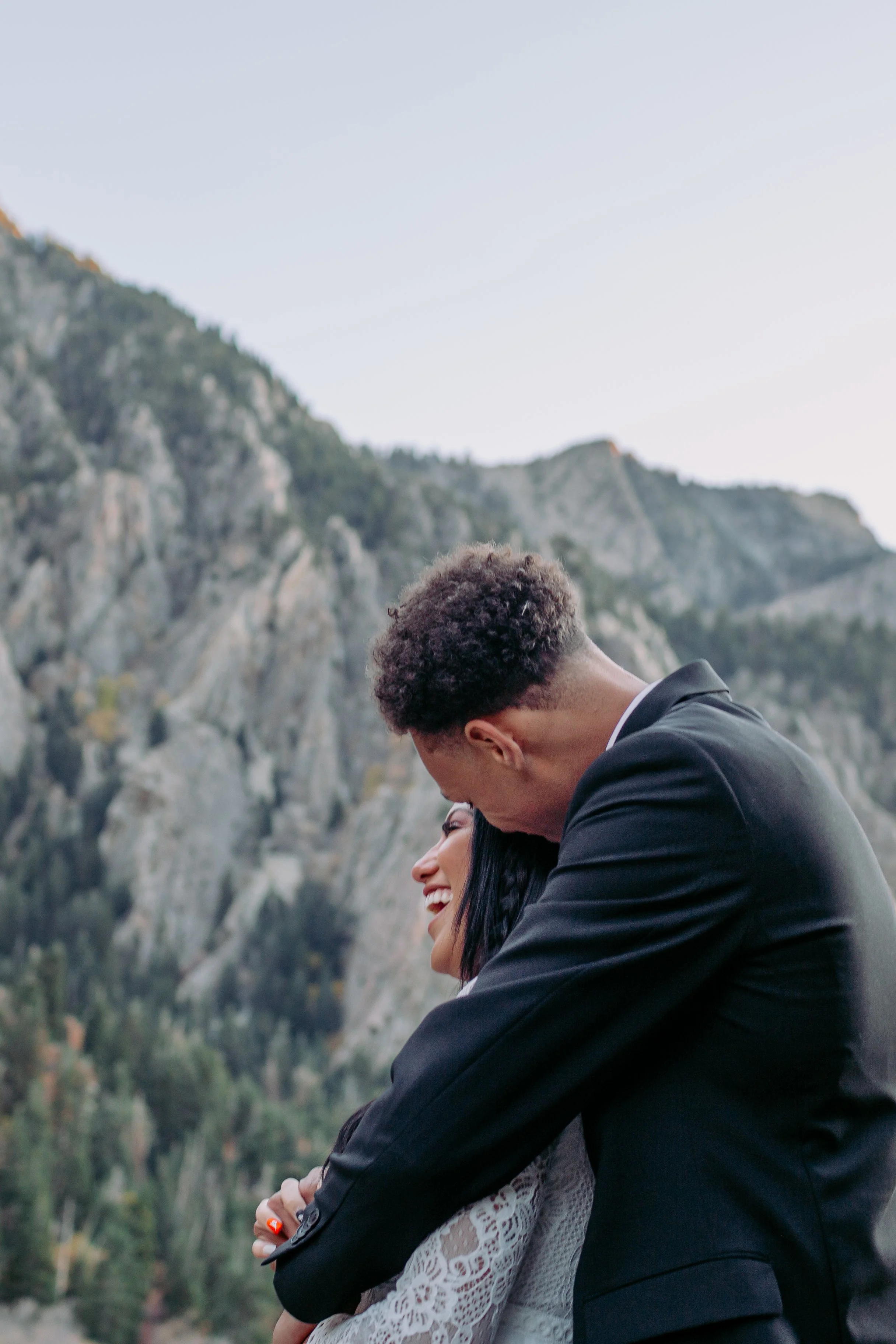 A couple embracing outdoors with forested mountains in the background, smiling and looking happy.