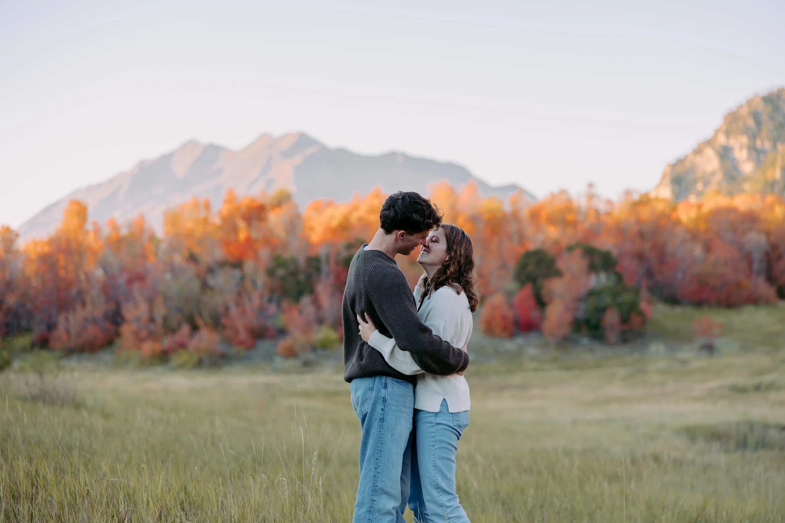 A couple embracing in a field with trees displaying fall colors and mountains in the background.