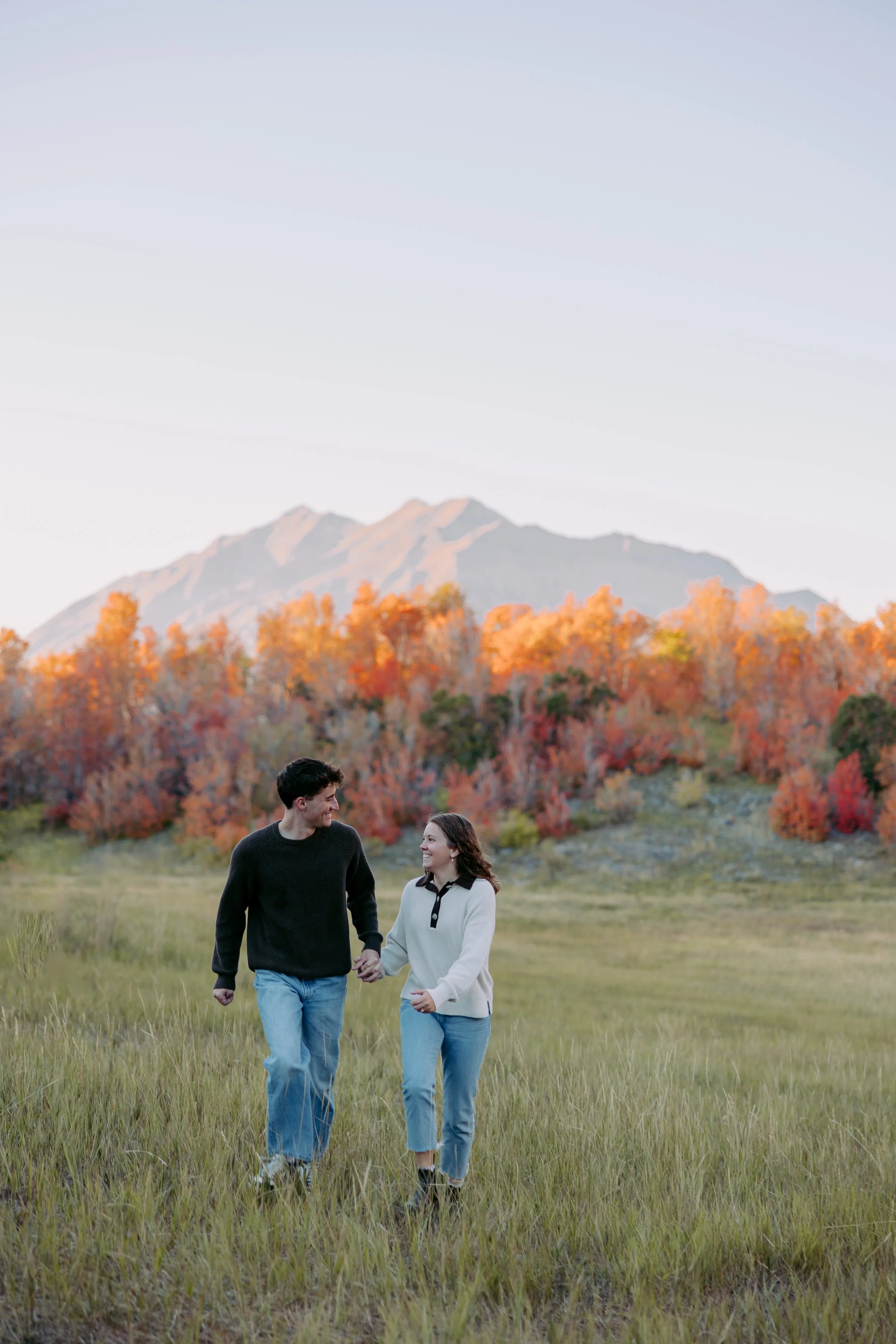 A young man and woman are walking hand-in-hand through a grassy field with autumn trees and a mountain in the background, during sunset.