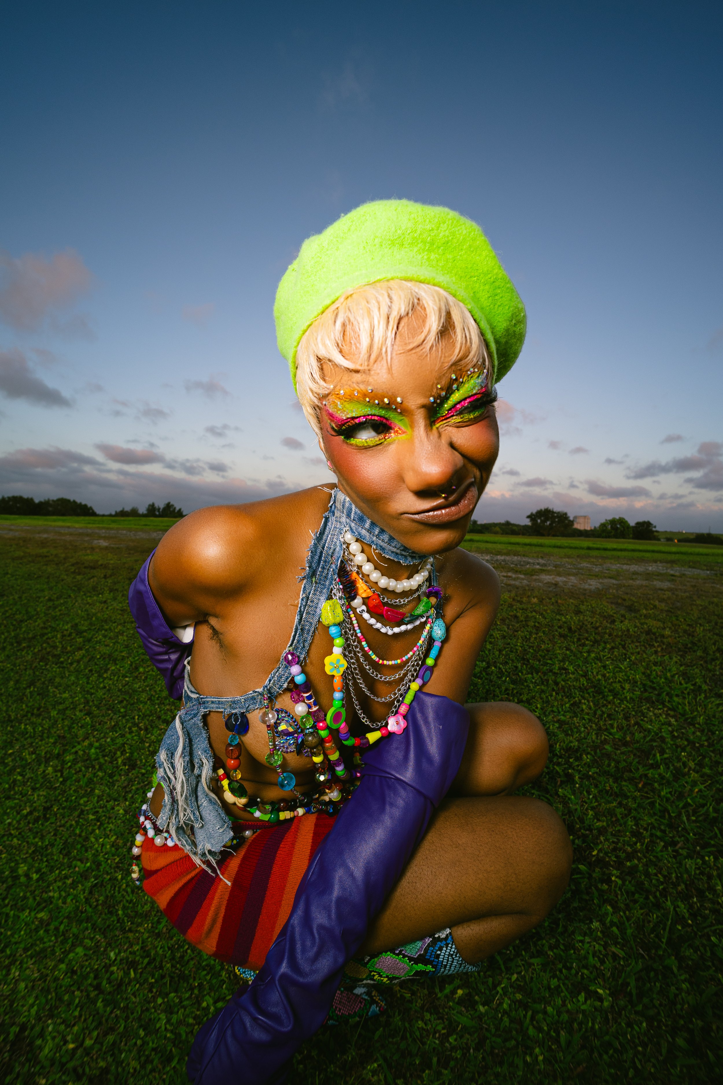 Person in colorful, eclectic outfit with bright makeup and accessories, wearing a green hat, kneeling outdoors on grass.