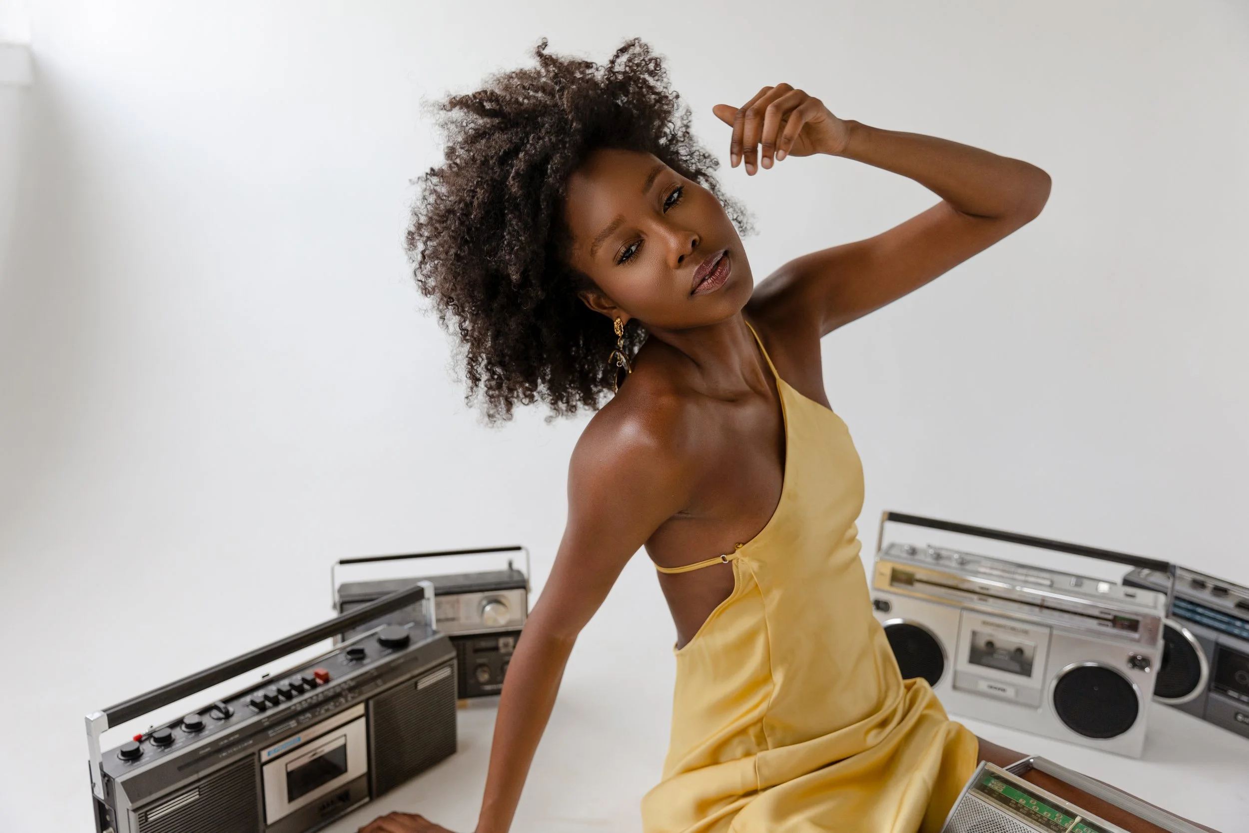 Woman in yellow dress sitting among vintage boomboxes on a white background.