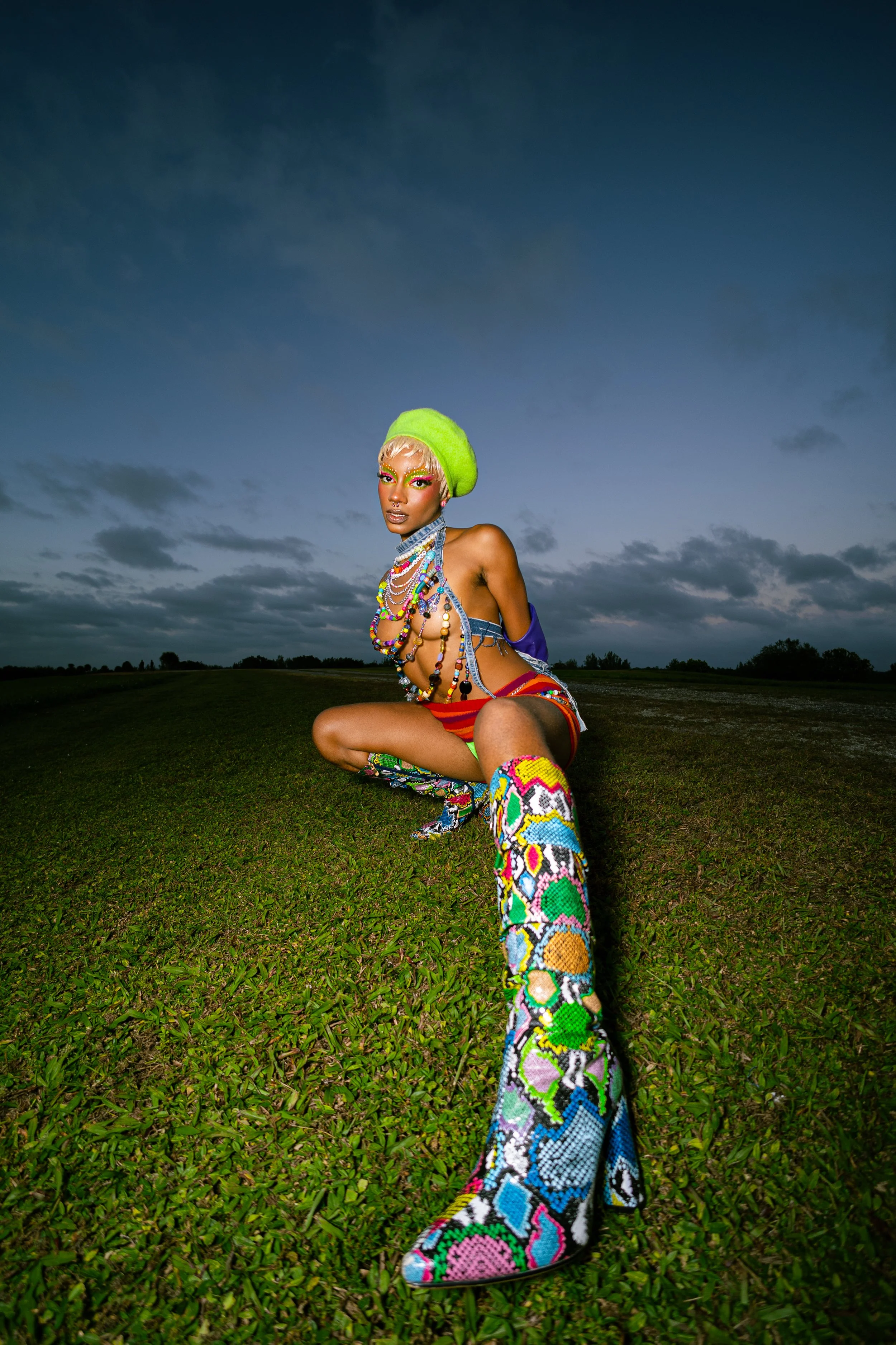Person wearing colorful outfit and boots, crouching on grass against a cloudy sky.