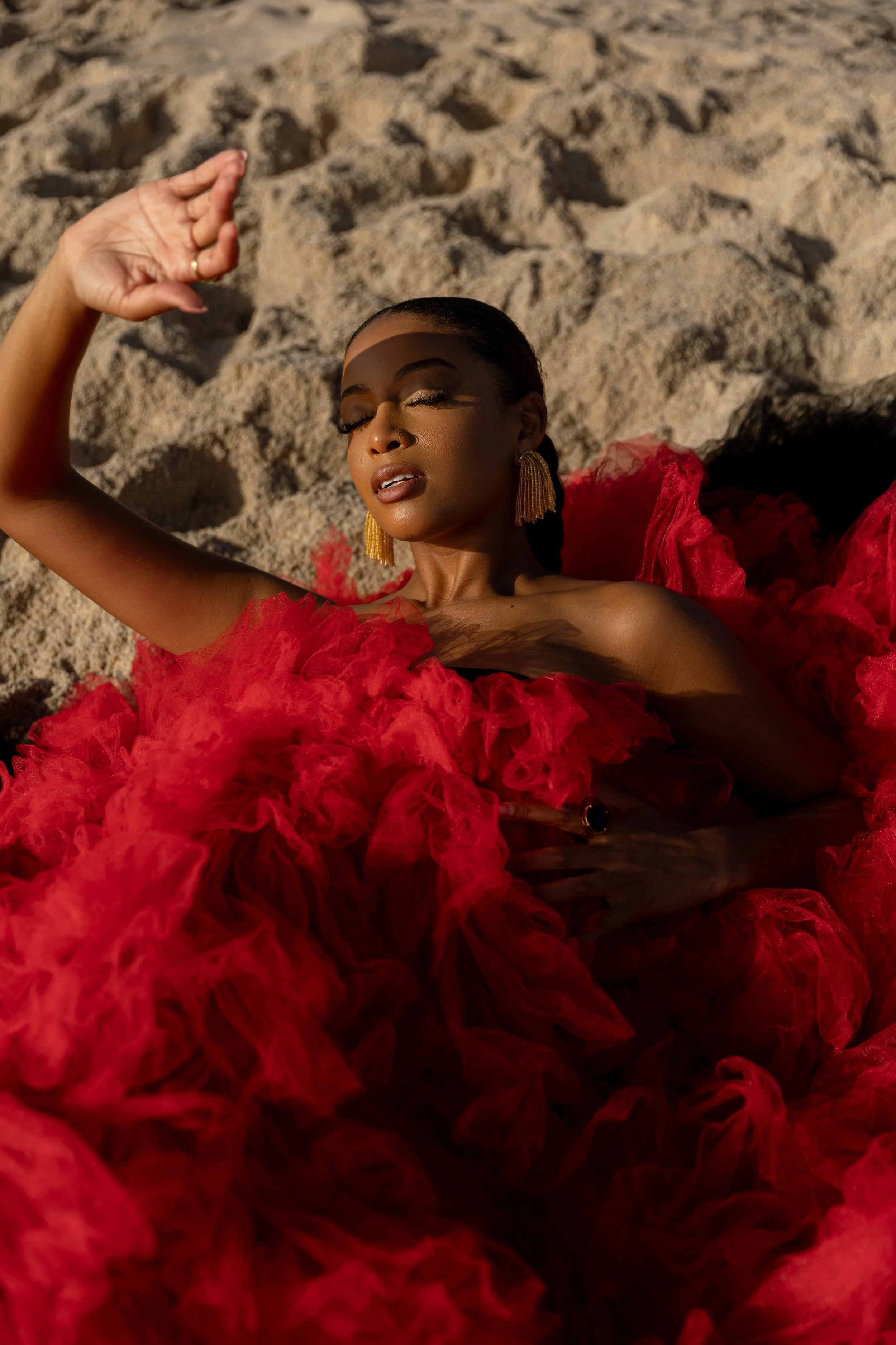 Woman in a voluminous red dress lying on sand, wearing gold tassel earrings, sunlight casting shadows on her face.