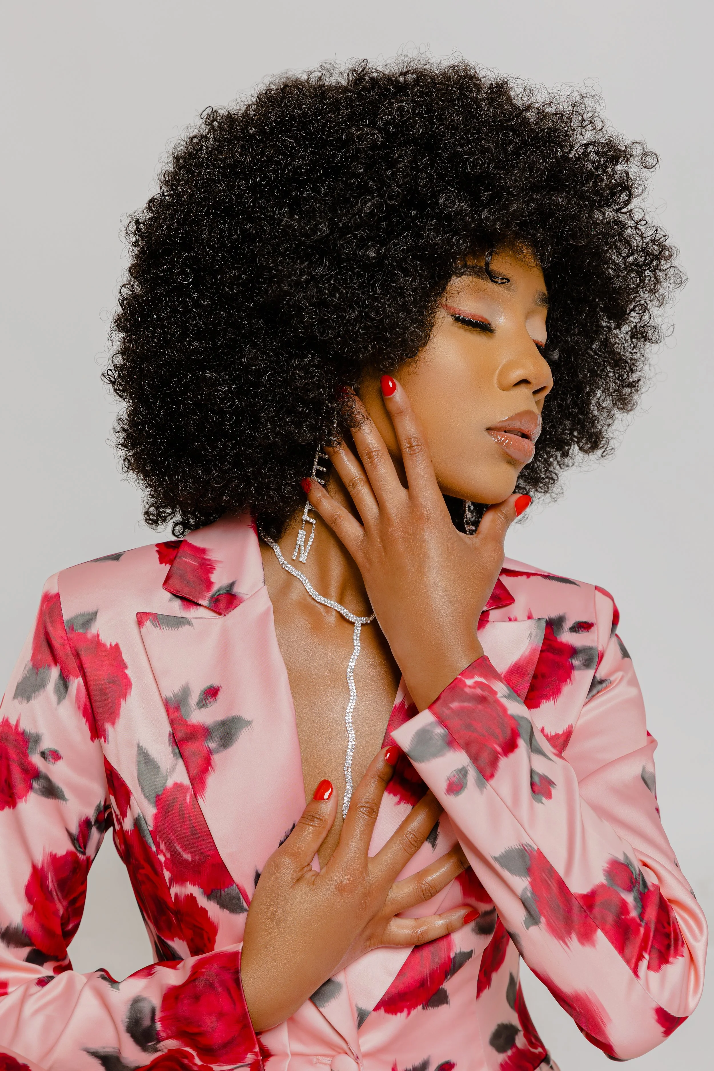 Person with curly hair in floral pink suit, wearing red nail polish and jewelry, posing with hands on face and chest. Minimalist, stylish look.