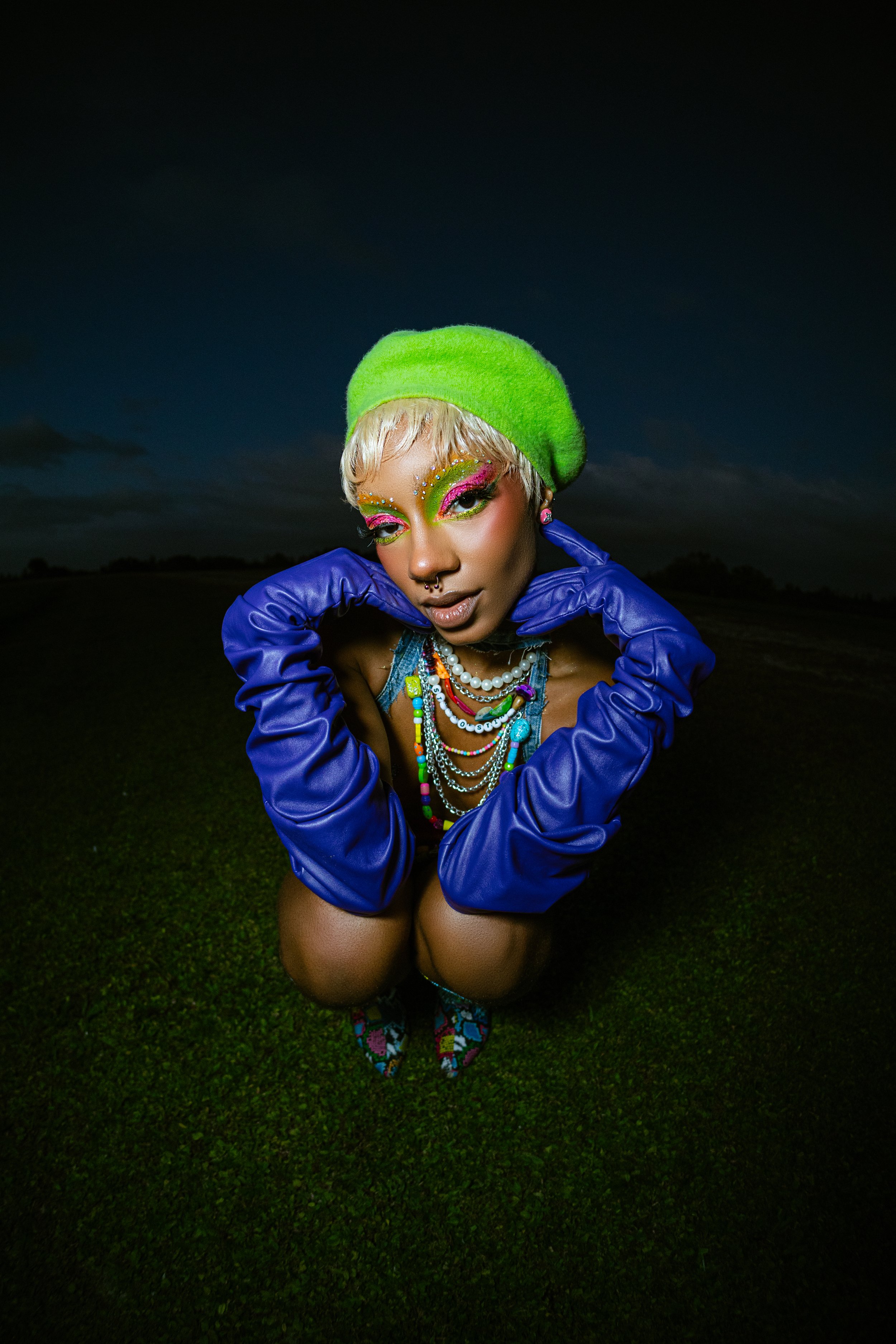 Person squatting on grass wearing bright blue gloves, a green beret, colorful makeup, and layered necklaces.