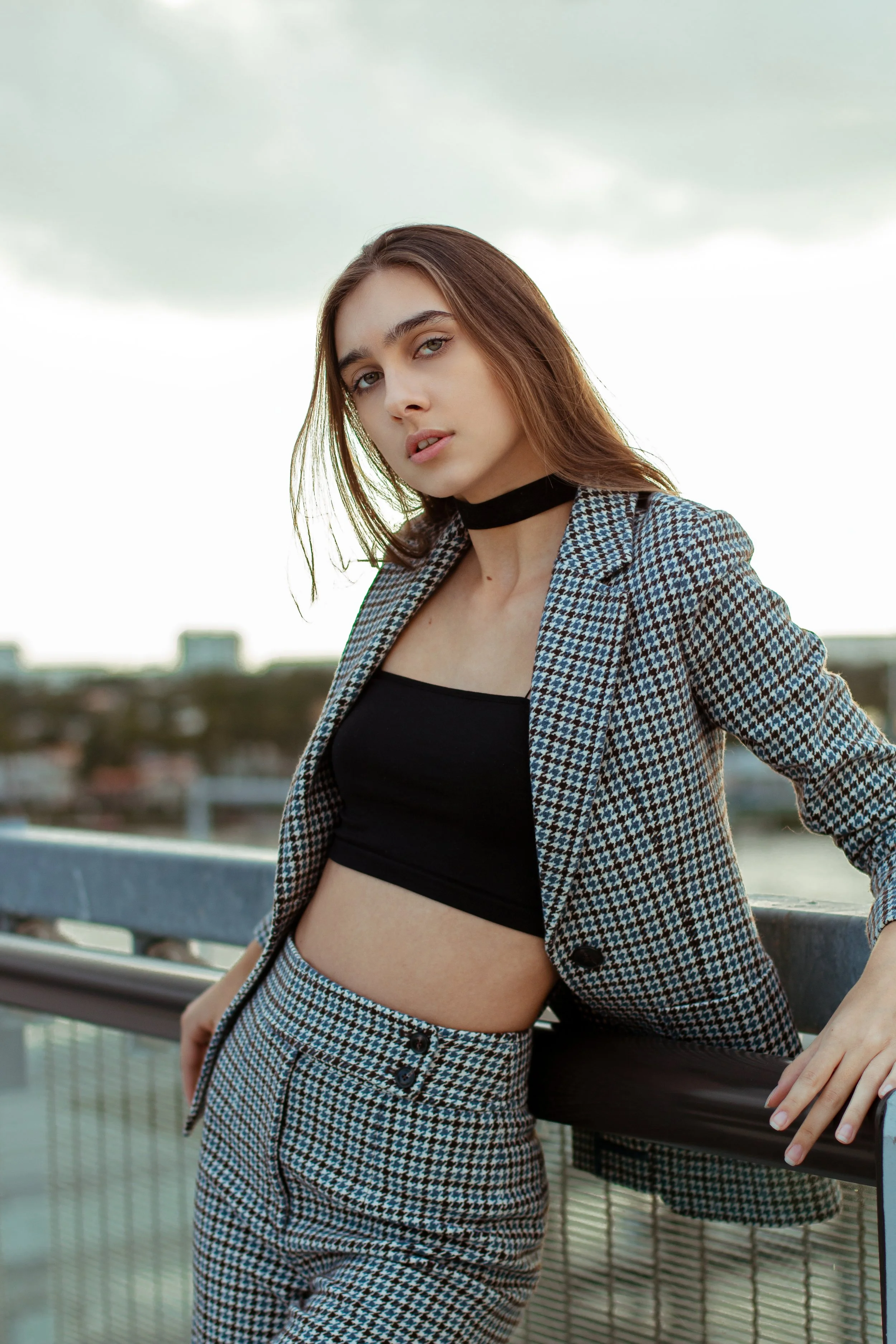 Young woman in fashionable checkered blazer and pants with black bandeau top, wearing a choker, leaning on a rail, outdoor setting.